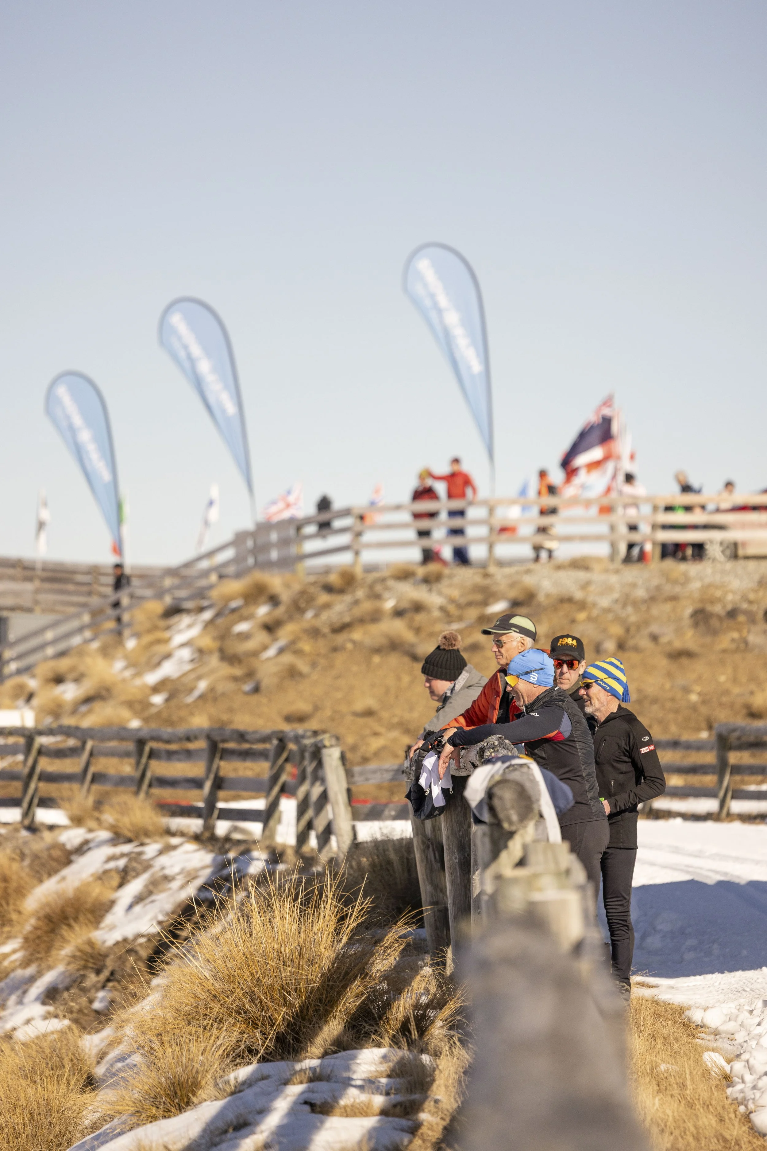 People walking along a snowy mountain trail with snow-covered ground and dry grass, wooden railings, flags, and blue banners in the background.