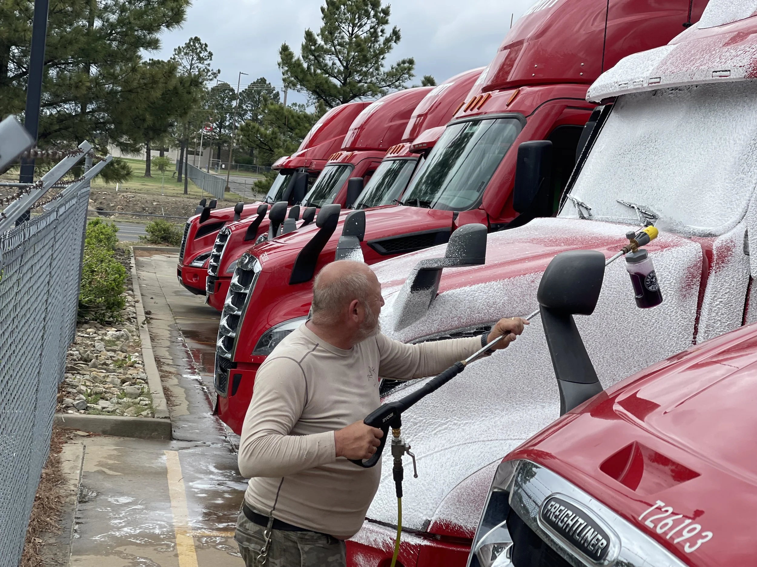 A man is pressure washing the front of a line of large red trucks in a parking lot.