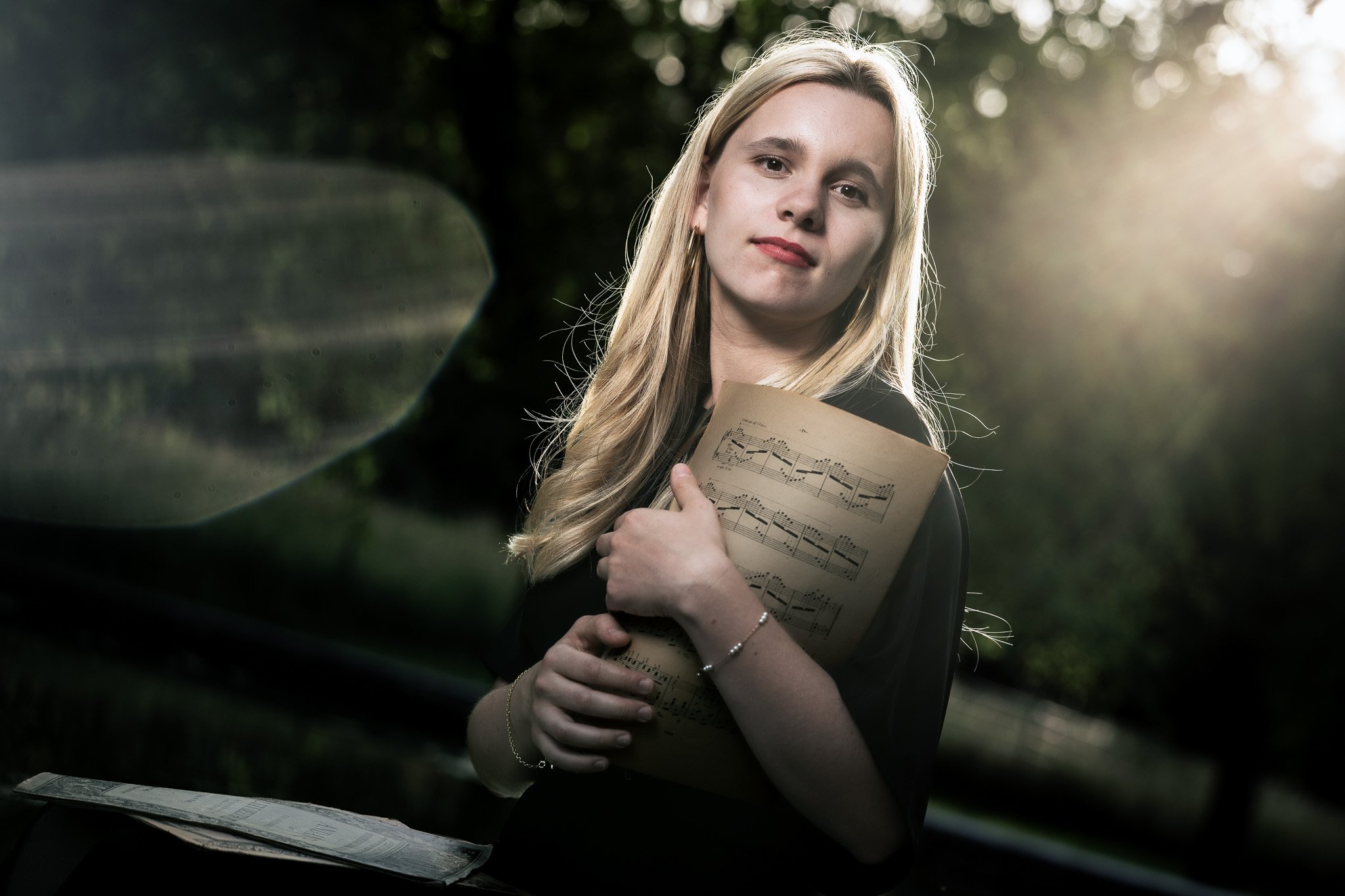 A woman with blonde hair holding sheet music, standing outdoors with sunlight filtering through trees.