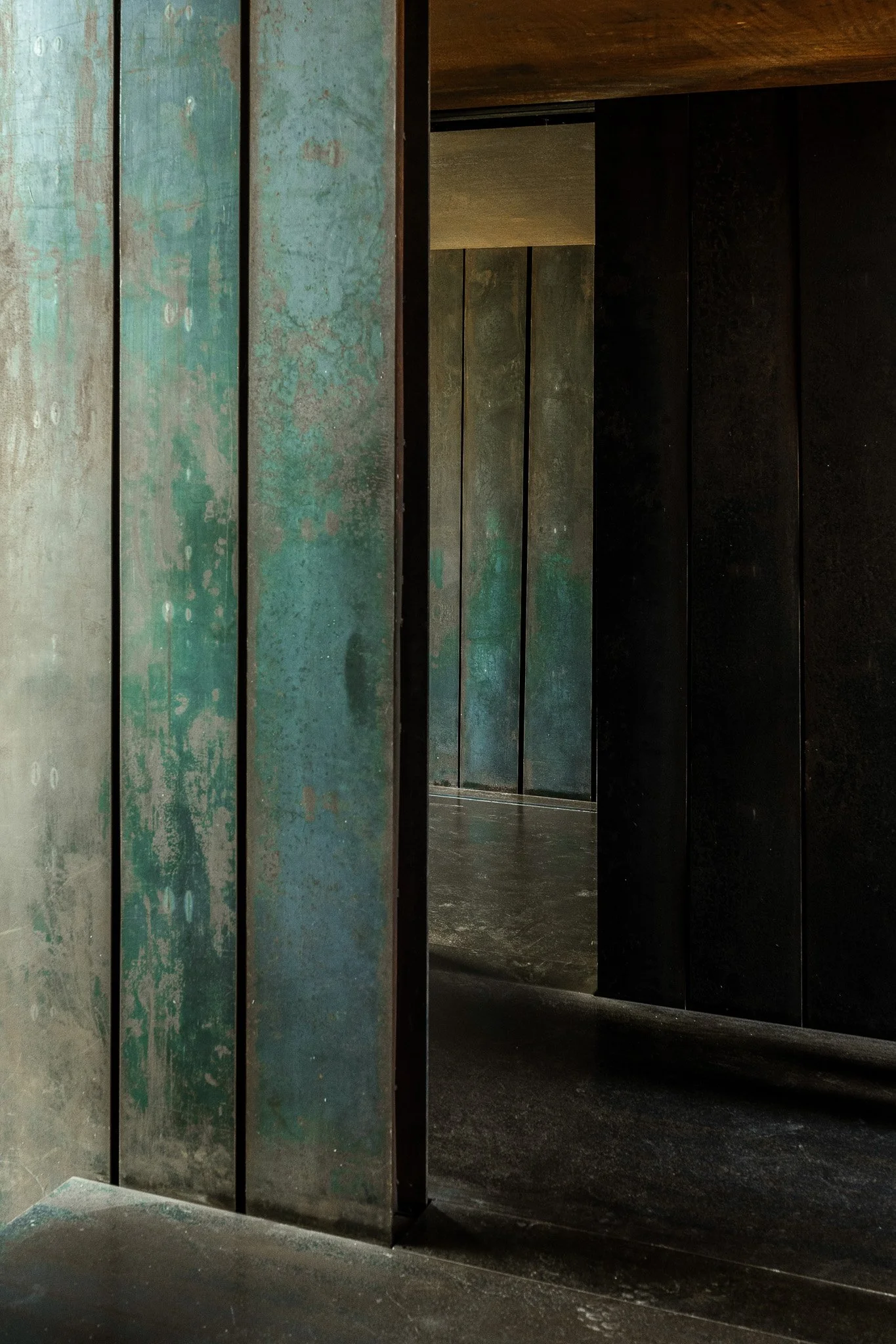 Close-up of aged, weathered metallic panels with a mix of greenish and rusty tones, separated by narrow black gaps, with a dark interior space visible behind.