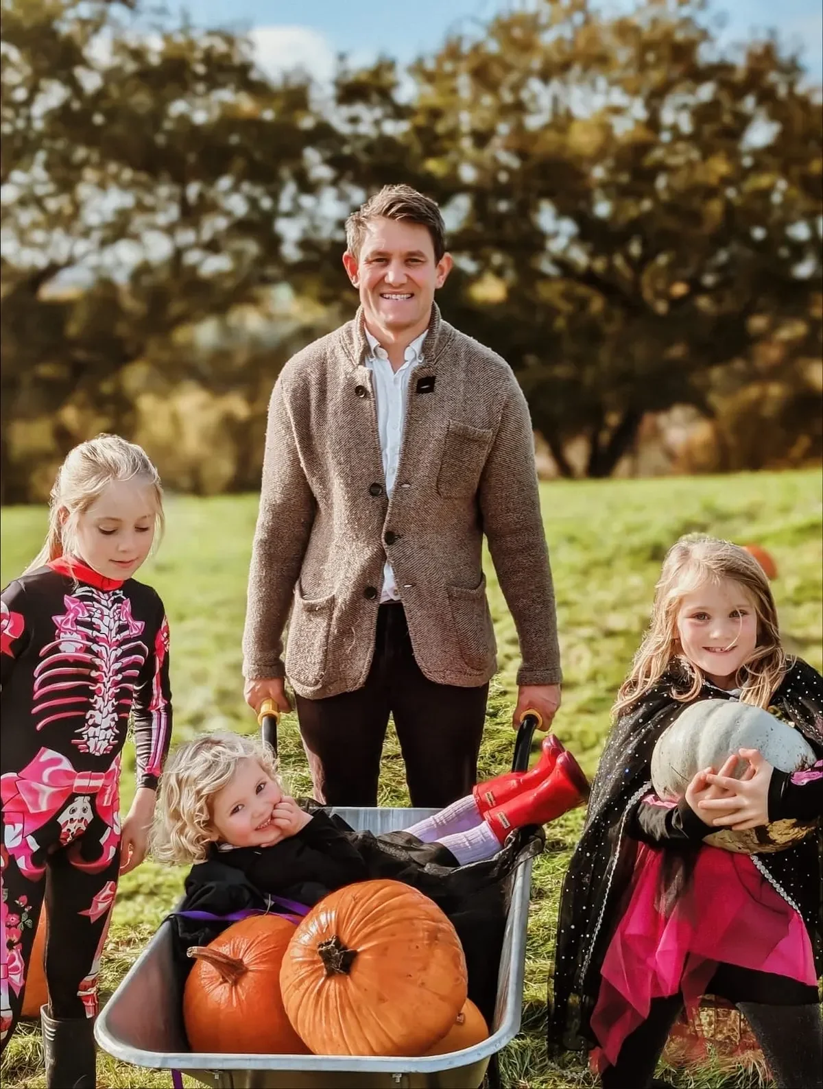 A man with children outdoors in a pumpkin patch, with pumpkins in a wheelbarrow, dressed in Halloween costumes.