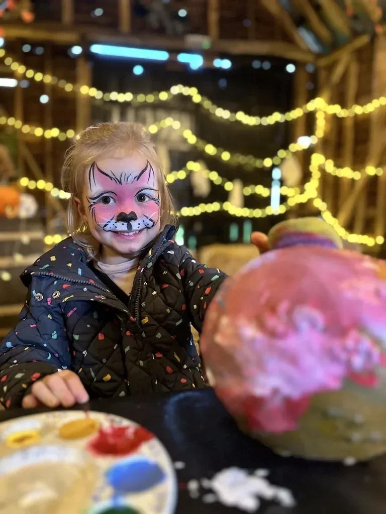 A young girl with face paint resembling a cat, smiling and sitting at a table with a painted pumpkin in the foreground. The background is decorated with yellow string lights, creating a festive atmosphere.