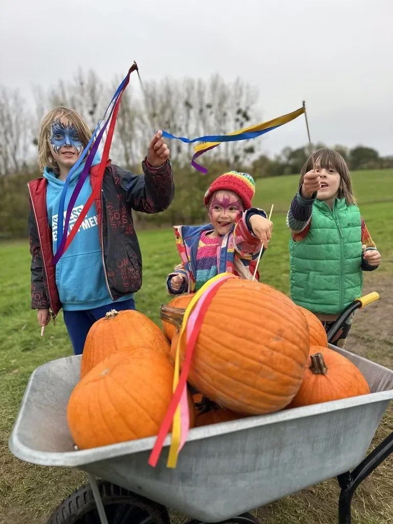 Three children with face paint and colorful clothing enjoying a pumpkin patch, surrounded by pumpkins in a wheelbarrow, outdoors on a cloudy day.