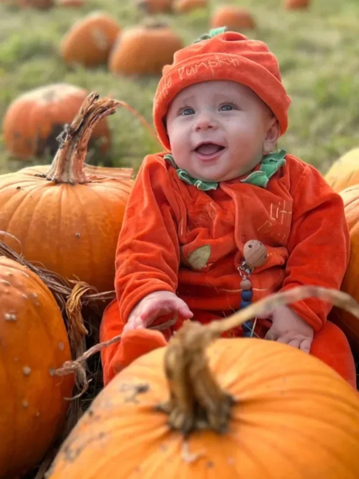A baby dressed in orange pumpkin-themed clothing sitting among pumpkins in a pumpkin patch, smiling and looking happy.