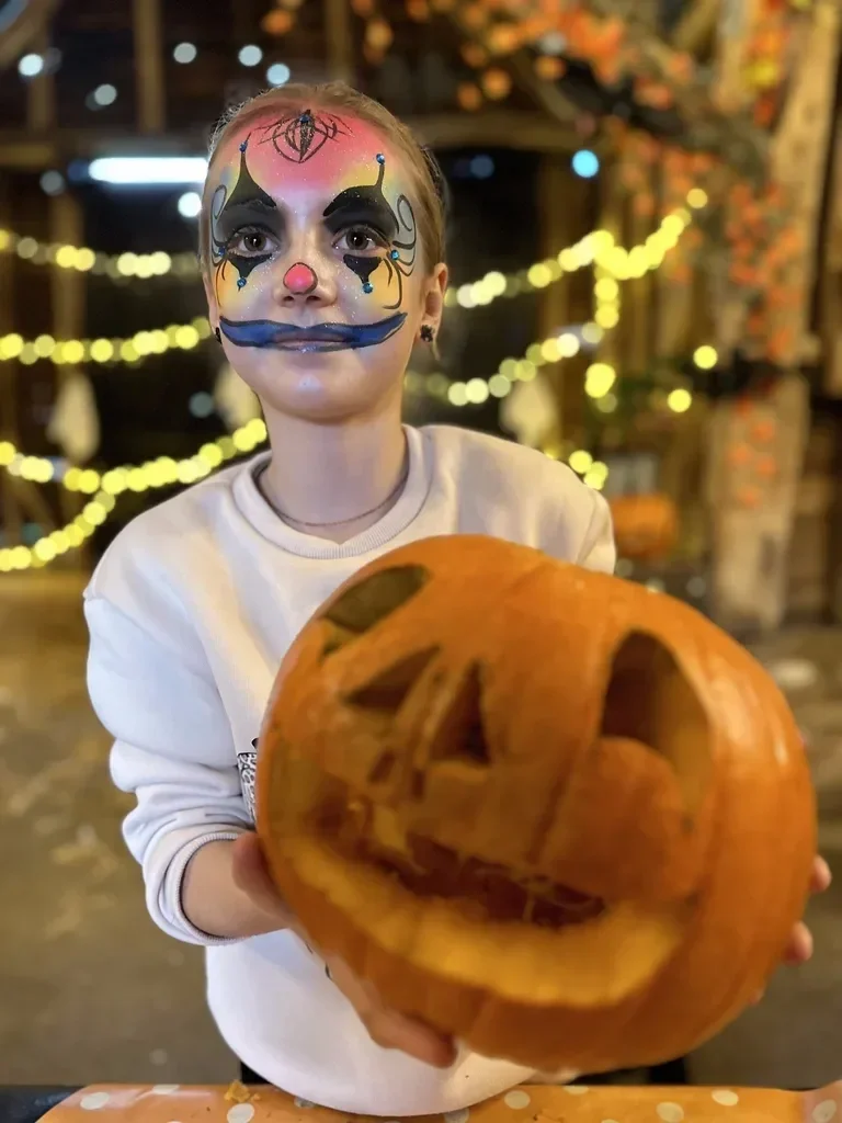 Girl with clown face paint holding a carved pumpkin with a Jack-o'-lantern face, standing in front of decorated background for Halloween.