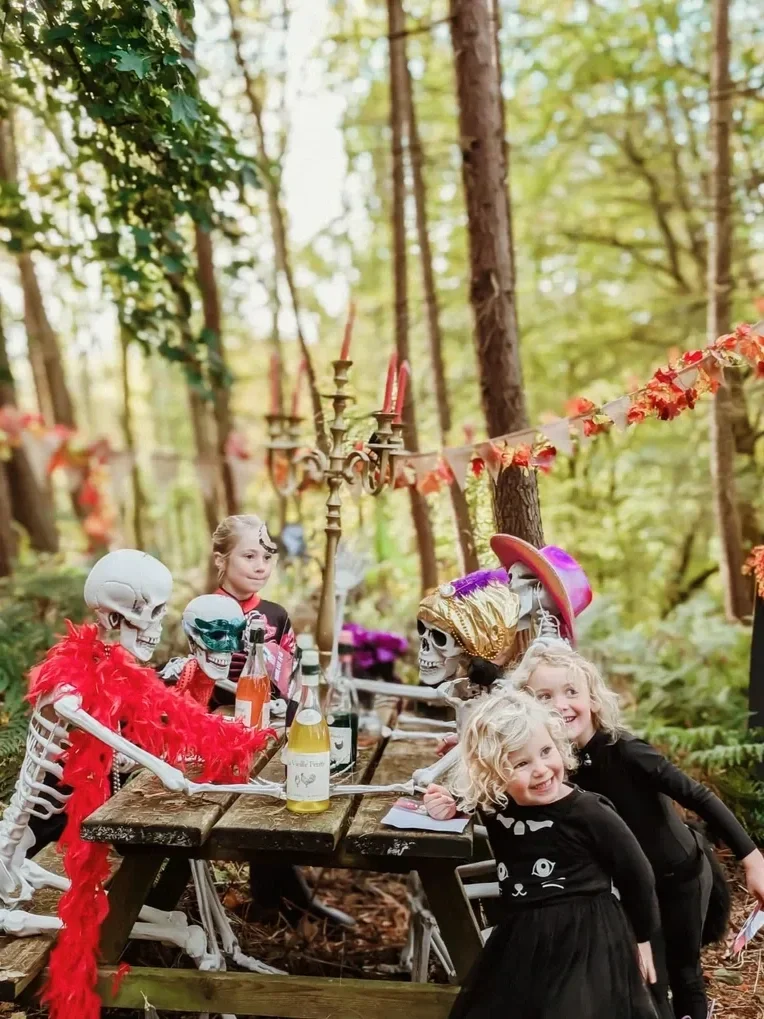 Three children dressed in black with ghost and skeleton decorations around a forest table, celebrating Halloween.
