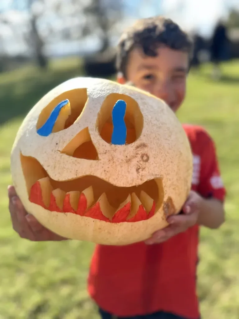 Child holding a carved pumpkin with a smiling face, decorated with blue and red paint, outdoors on a sunny day.