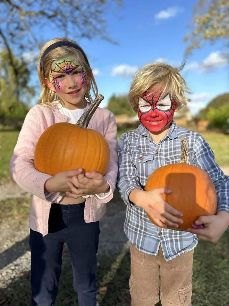 Two children with face paint holding pumpkins outdoors on a sunny day.