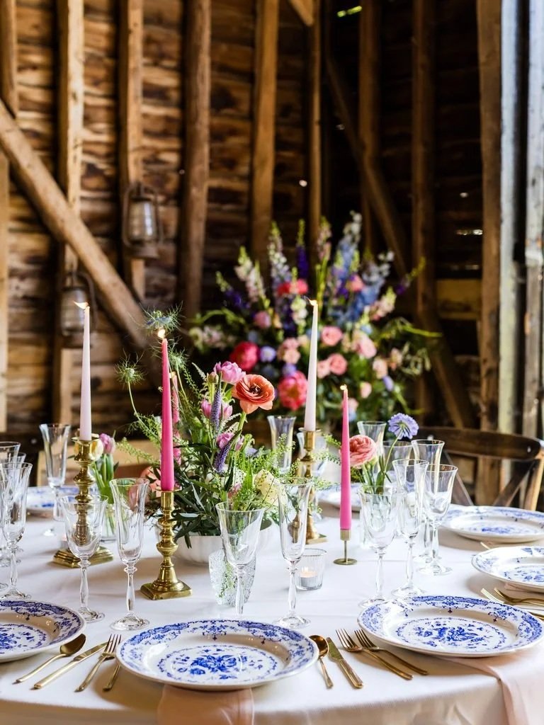 Table set for a formal event with floral centerpiece, pink and blue candles, gold flatware, and blue and white patterned plates in a rustic wooden barn.