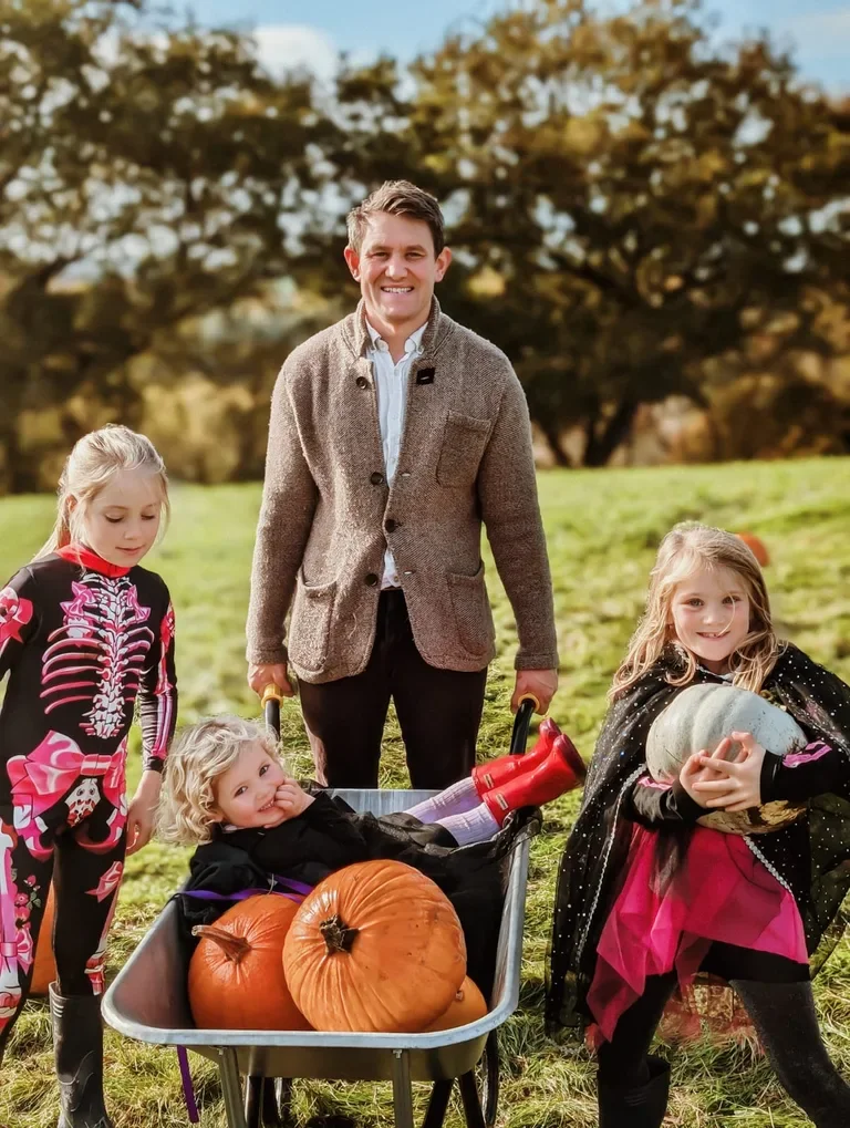 A man and three young girls outdoors on aan Autumnday, with pumpkins, some of the children dressed in Halloween costumes.