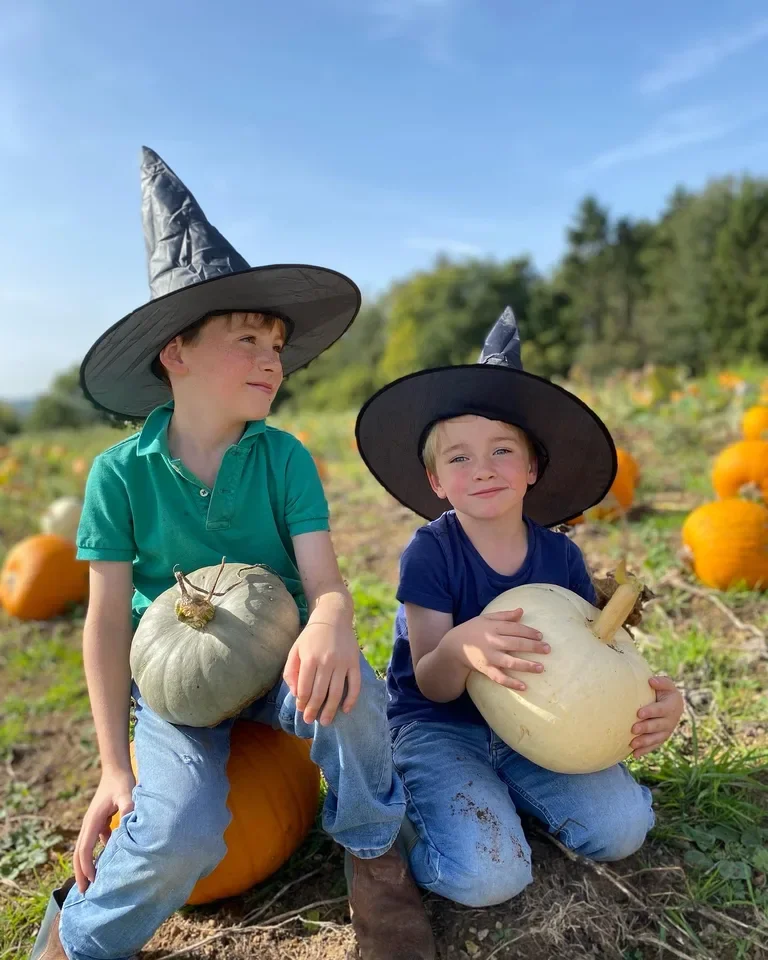 Two young boys wearing witch hats sitting on pumpkins in a pumpkin patch, holding white pumpkins, with other pumpkins scattered around, under a clear blue sky.