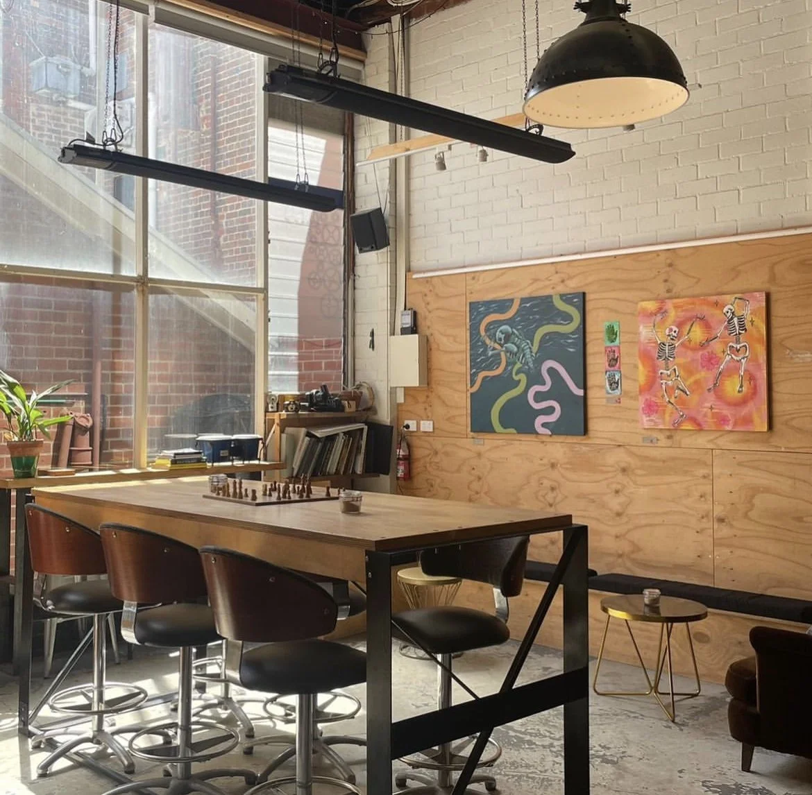 Interior of a modern art studio with a long wooden table, black chairs, large window, potted plant, colorful artwork on the wall, and hanging industrial lights.
