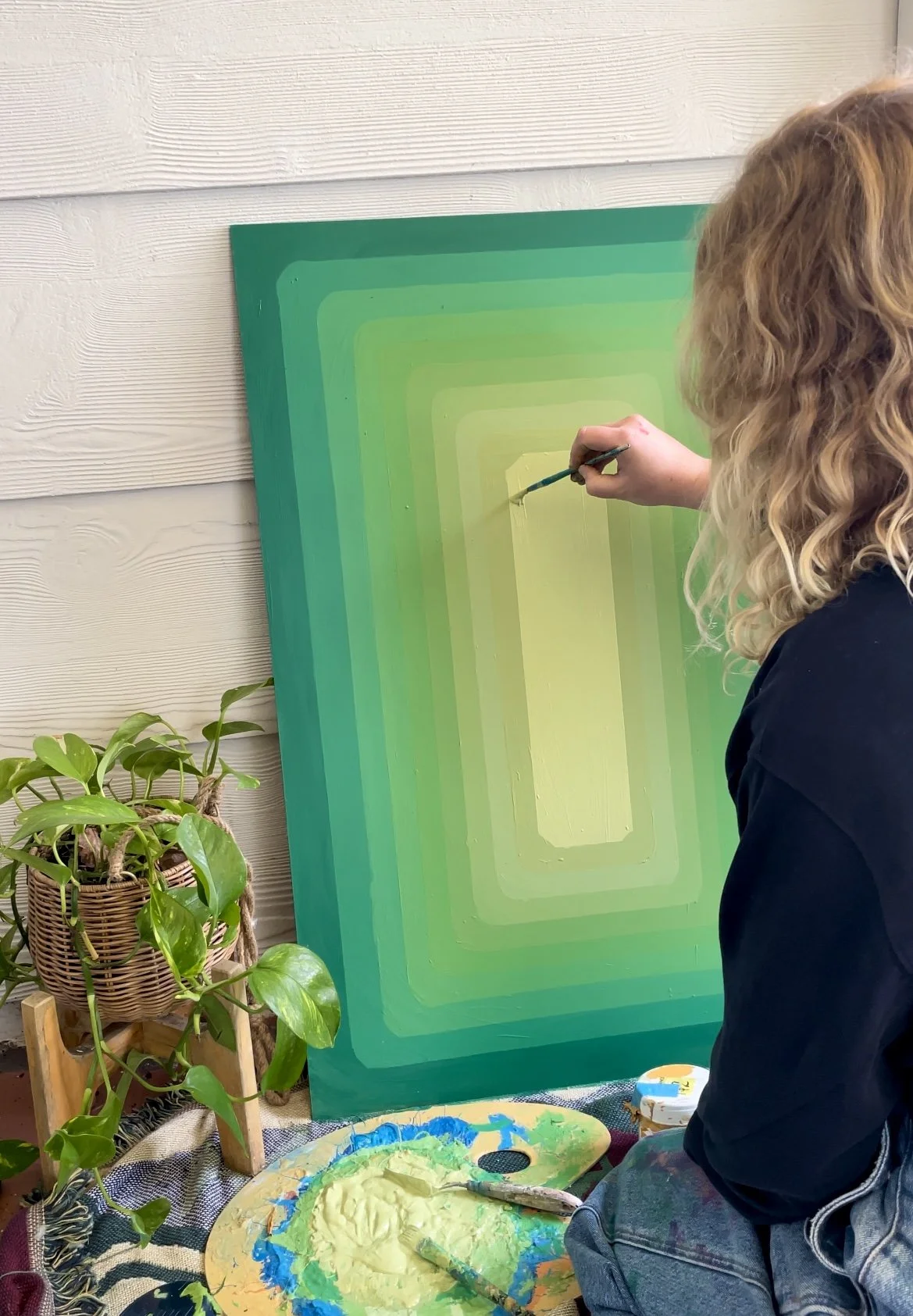 A woman painting a geometric abstract artwork with gradients of green and yellow on a large canvas, sitting next to a potted plant.