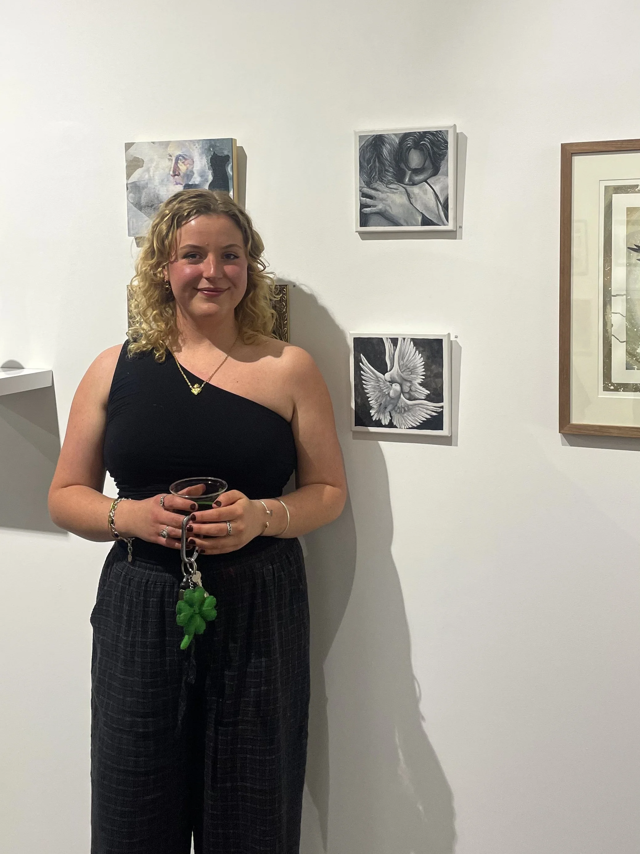 A woman with curly blonde hair wearing a one-shoulder black top and black checkered pants, standing in an art gallery, holding a drink with a green keychain.