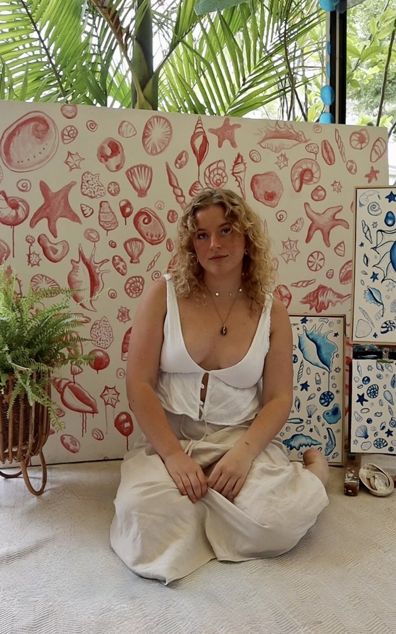 A woman with curly blonde hair sits on the floor in front of a beach-themed mural with red and blue sea creatures and shells, surrounded by green plants and artwork.