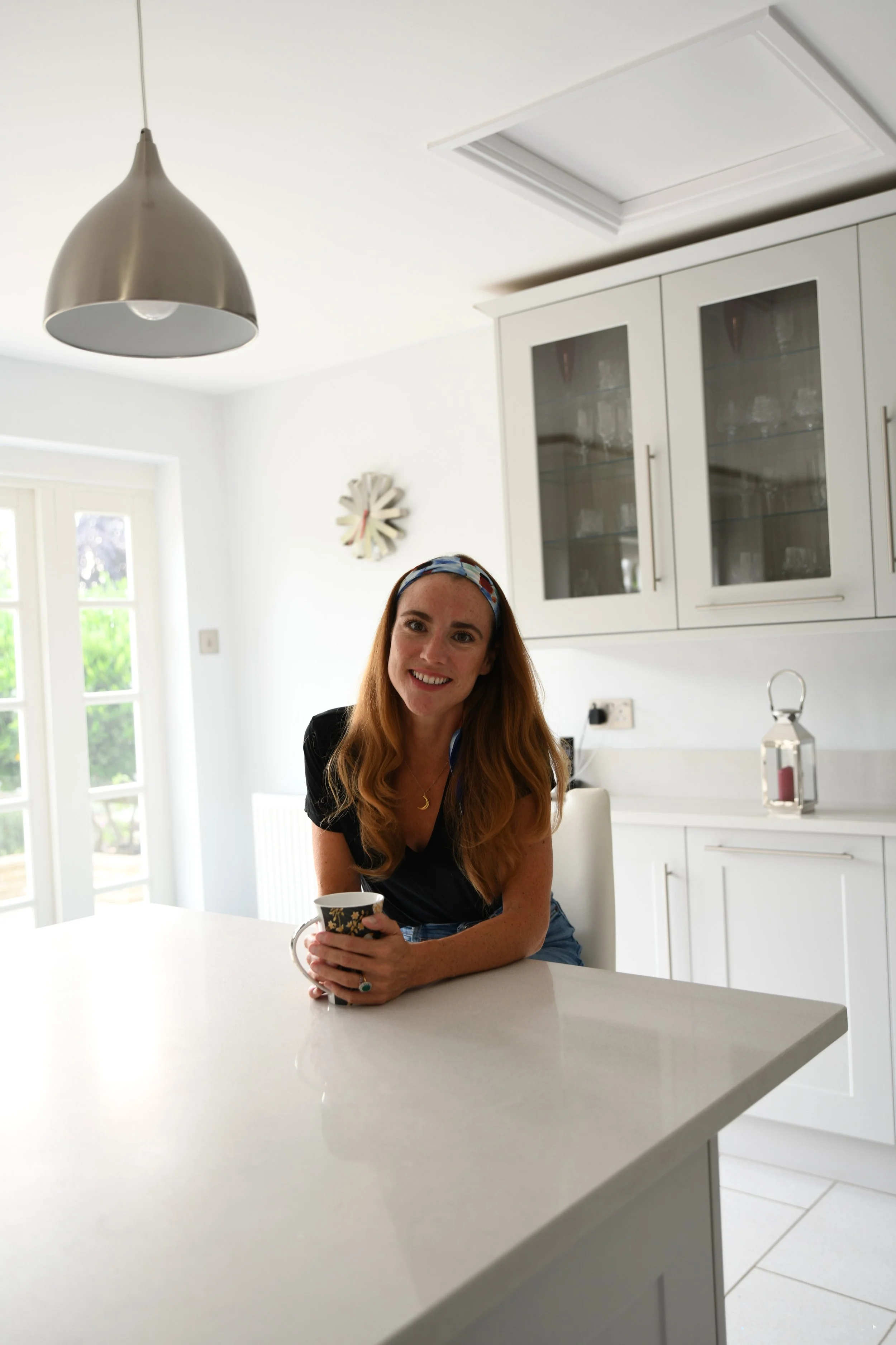 Woman standing in a modern kitchen, holding a cup, with white cabinets and glass doors in the background.
