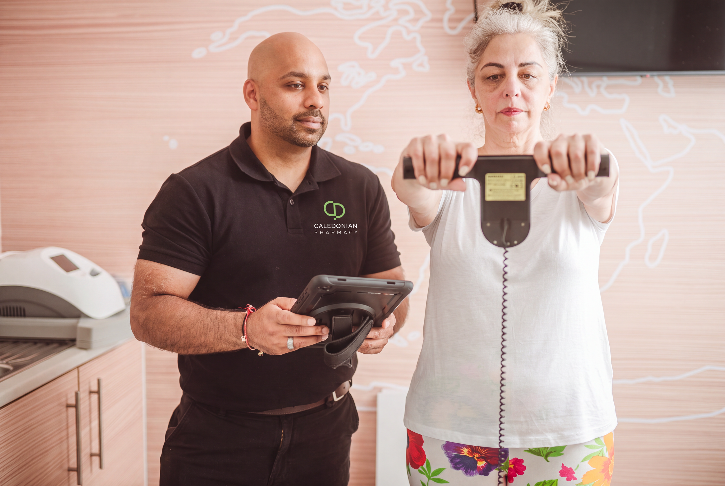 A male healthcare worker in a shirt with a 'Caledonian Pharmacy' logo assisting a smiling woman with gray hair, who is wearing a white top and colorful floral pants, as she gets weighed in a consultation.