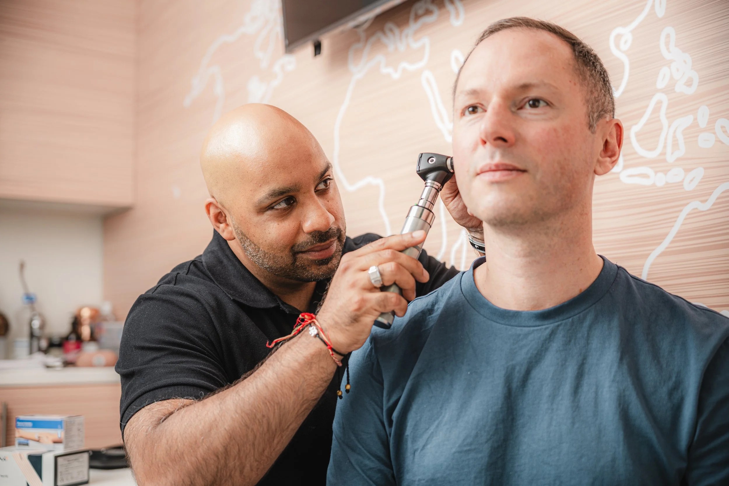 A man receiving an ear examination from a healthcare professional in a medical office with world map and lines on the wall in the background.