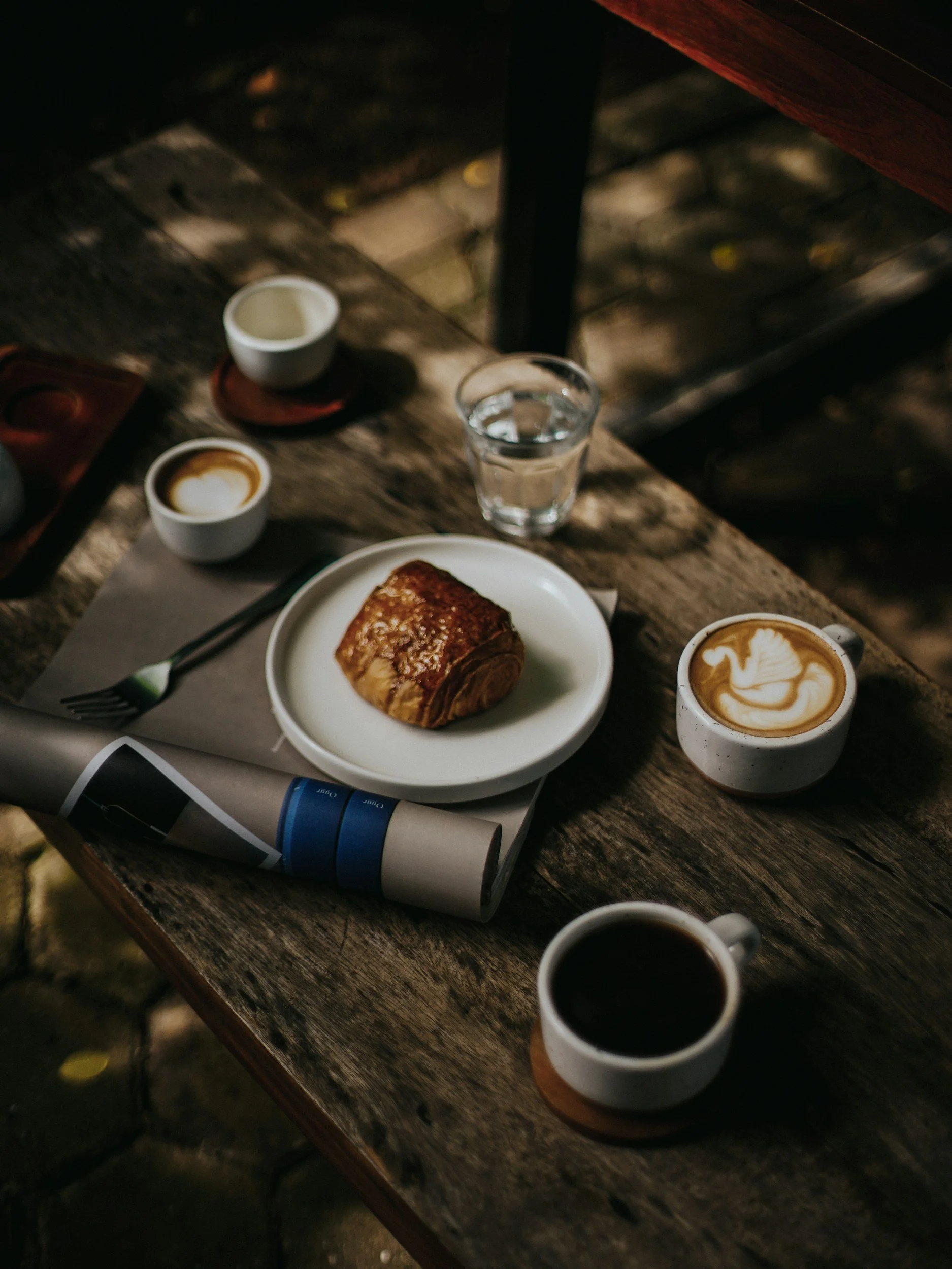 A rustic wooden table set with cups of coffee, a glass of water, and a slice of toast or pastry.