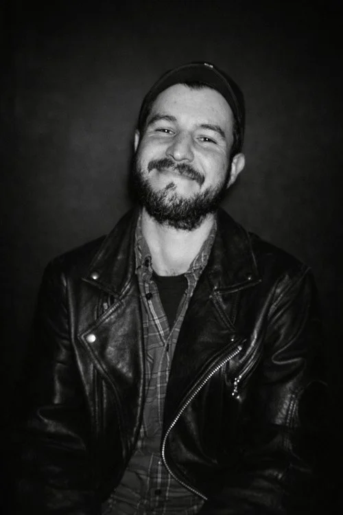 A headshot of Patrick, a man with a beard and a mustache smiling at the camera in black and white.