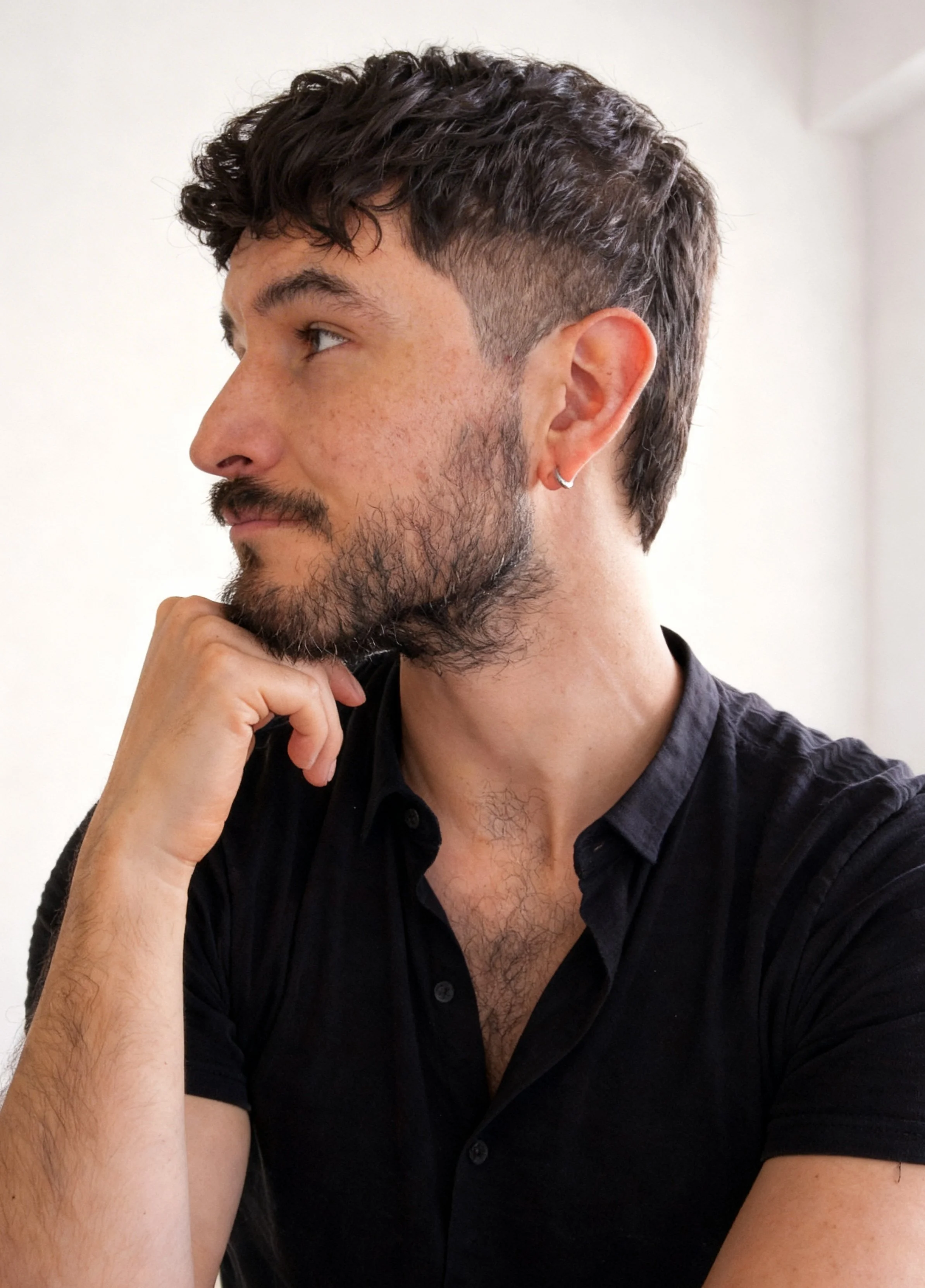 A man with dark, curly hair, a beard, and a mustache, wearing a black collared shirt, resting his chin on his hand, looking to the left, with a plain white wall background.