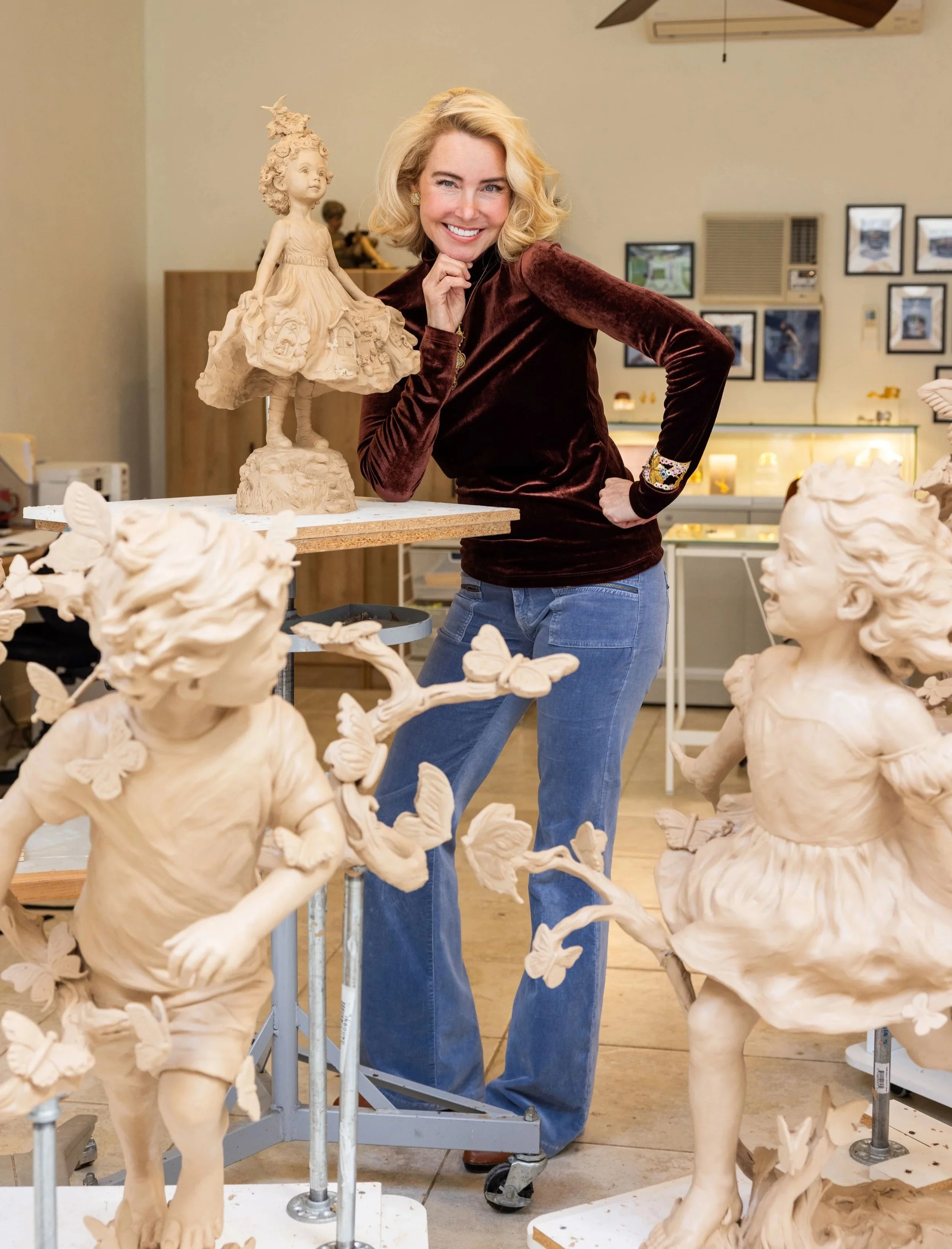 A woman with blonde hair smiling and posing next to clay sculptures of children with butterflies in an art studio.