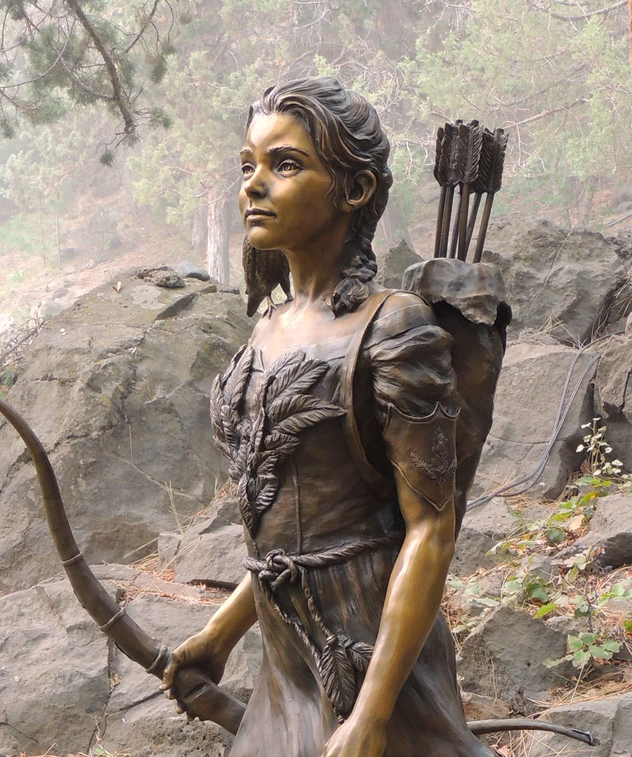 Bronze sculpture of a young Native American woman with a bow in her hand, wearing traditional clothing and carrying a quiver of arrows on her back, situated outdoors among rocks and trees.