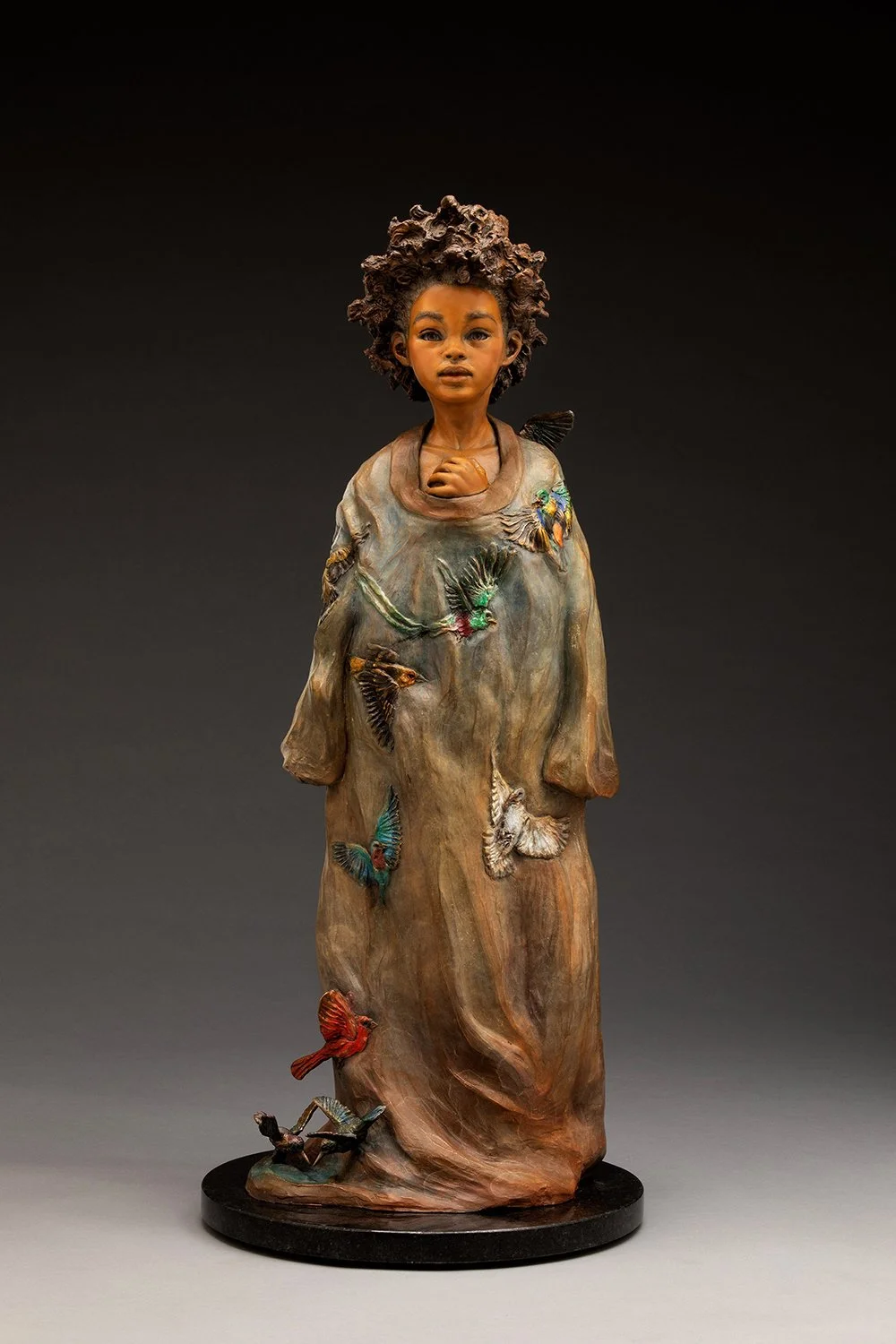 A sculpture of a young girl with curly hair wearing a brown robe decorated with colorful birds and butterflies, standing on a round black base.