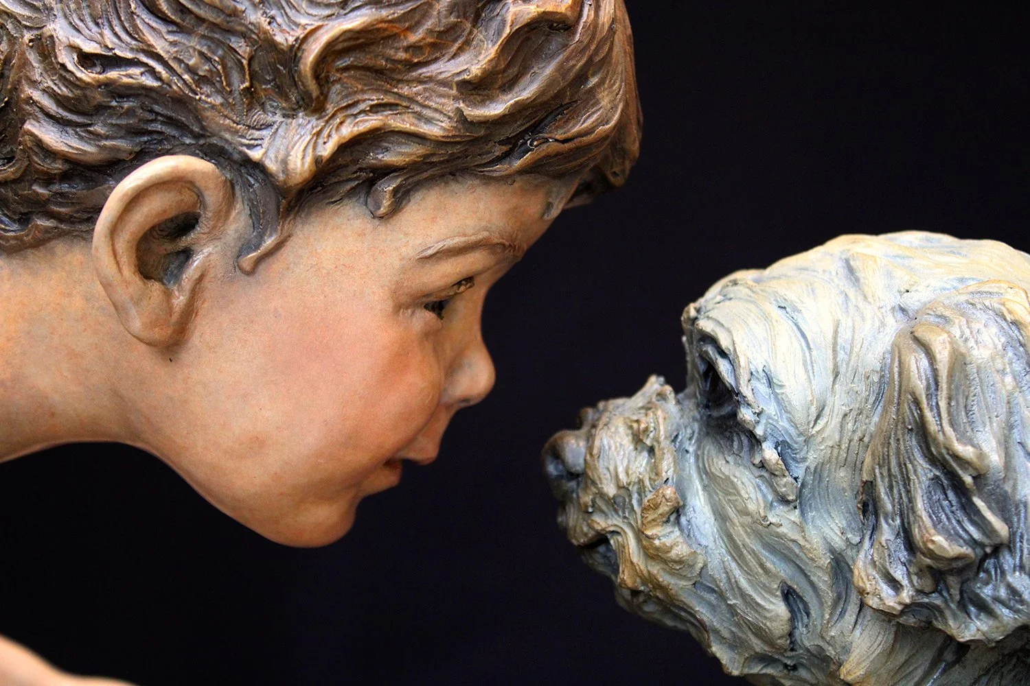 Close-up of a sculpture of a young boy's face with detailed hair and profile, facing a sculpture of a dog's face with textured fur, both set against a black background.