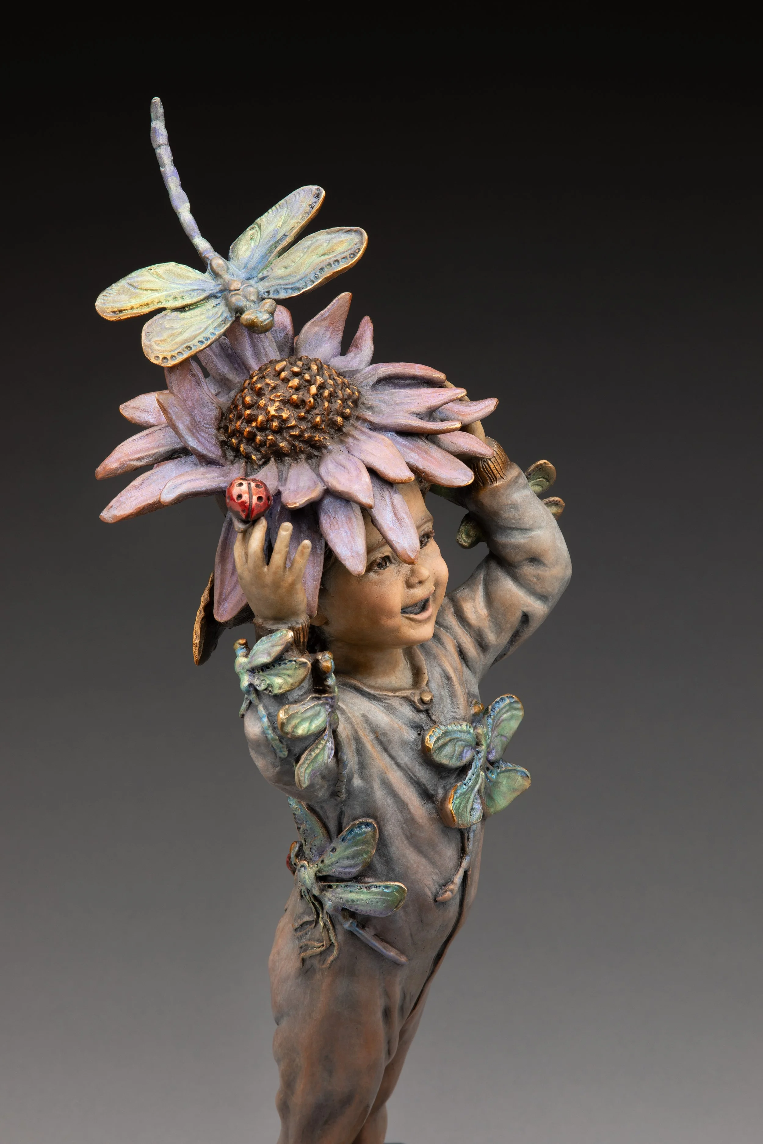 A sculpture of a young girl holding a large sunflower on her shoulder, with a dragonfly perched on the sunflower and a ladybug on her hand, set against a plain dark background.