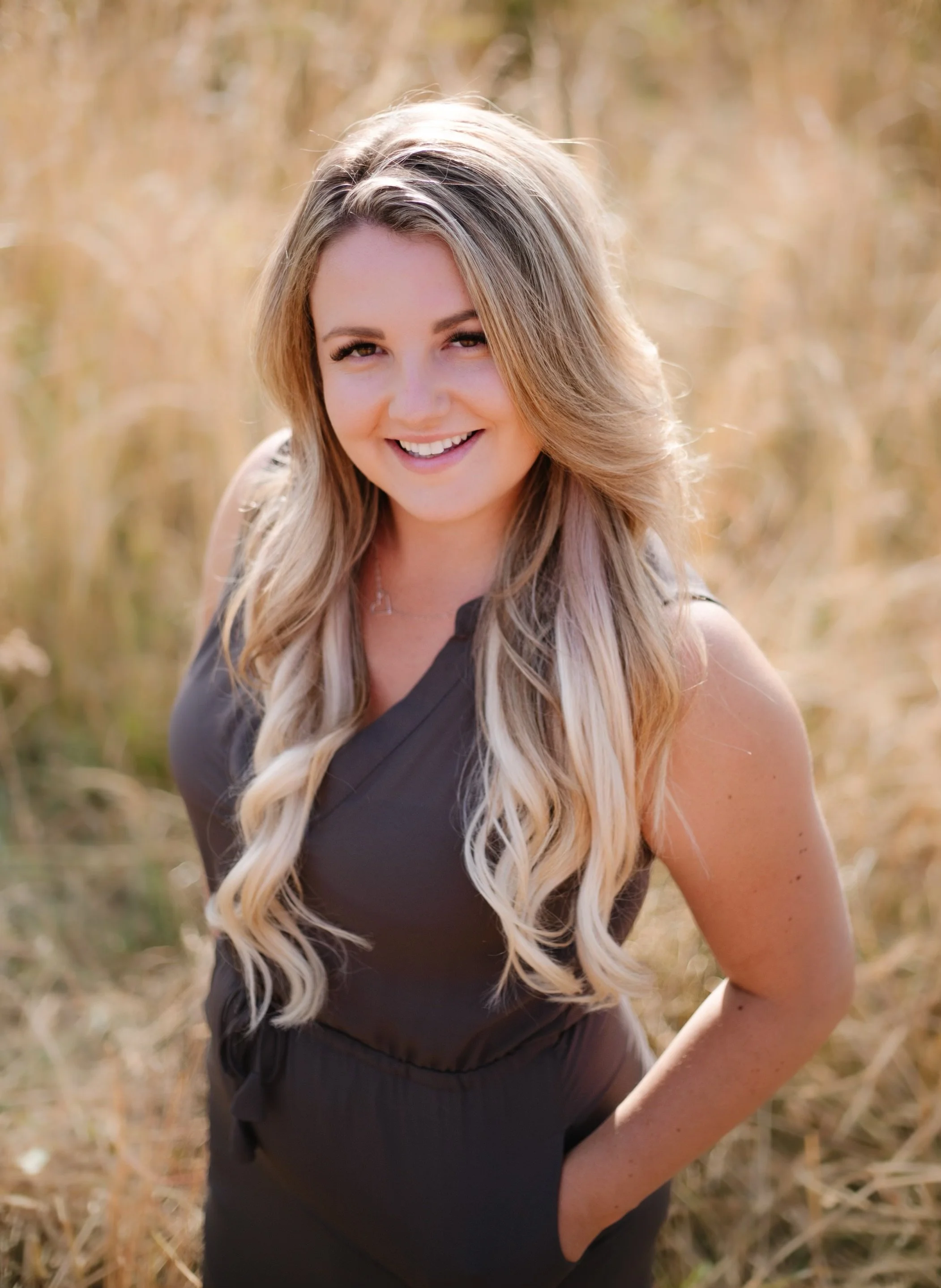 Smiling woman with long blonde hair standing in a field of tall grass.
