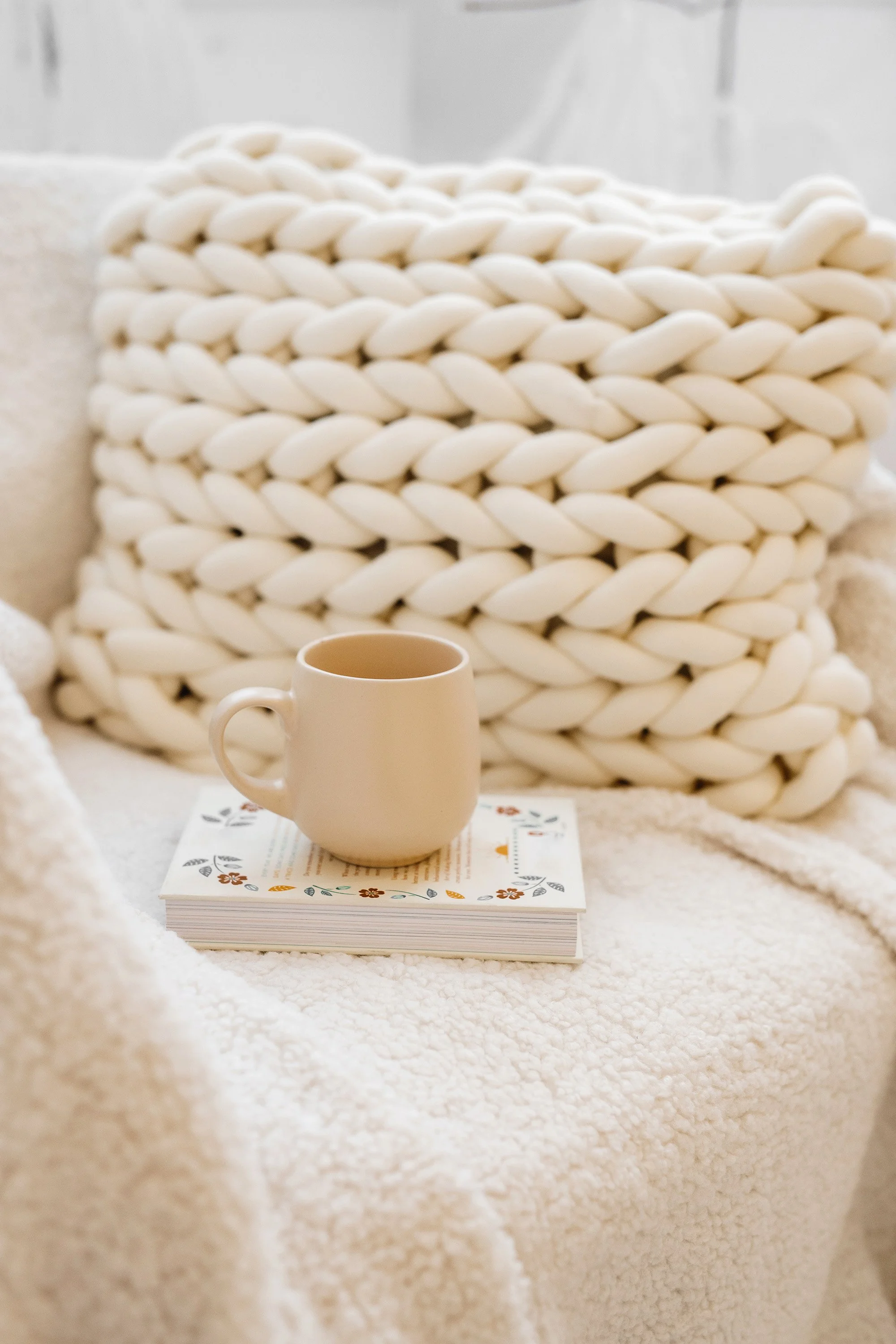 Cozy setting with a beige ceramic mug on a book, featuring a knitted pillow and a fluffy blanket.