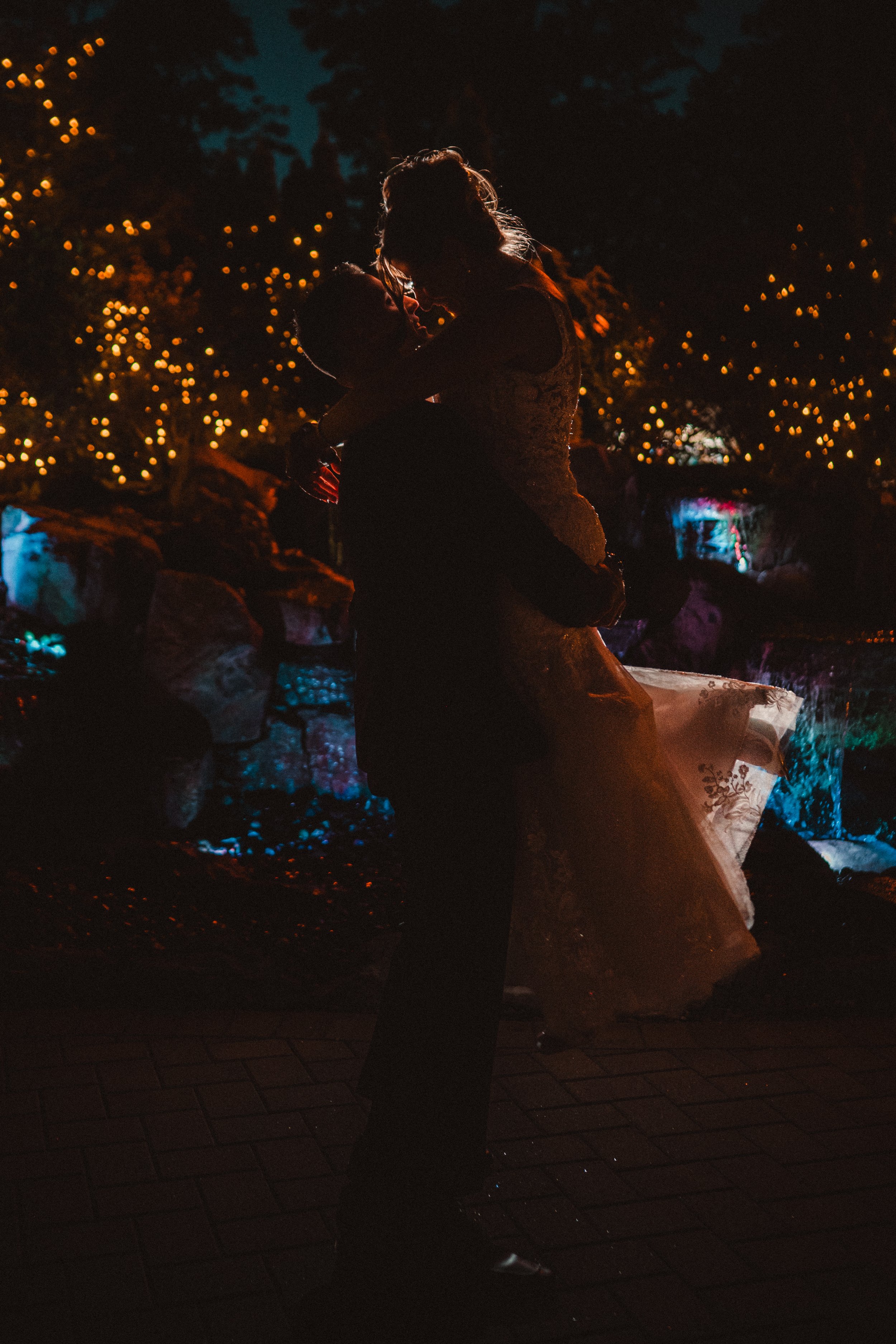 Silhouette of a couple dancing outdoors at night with string lights and colorful water features in the background.