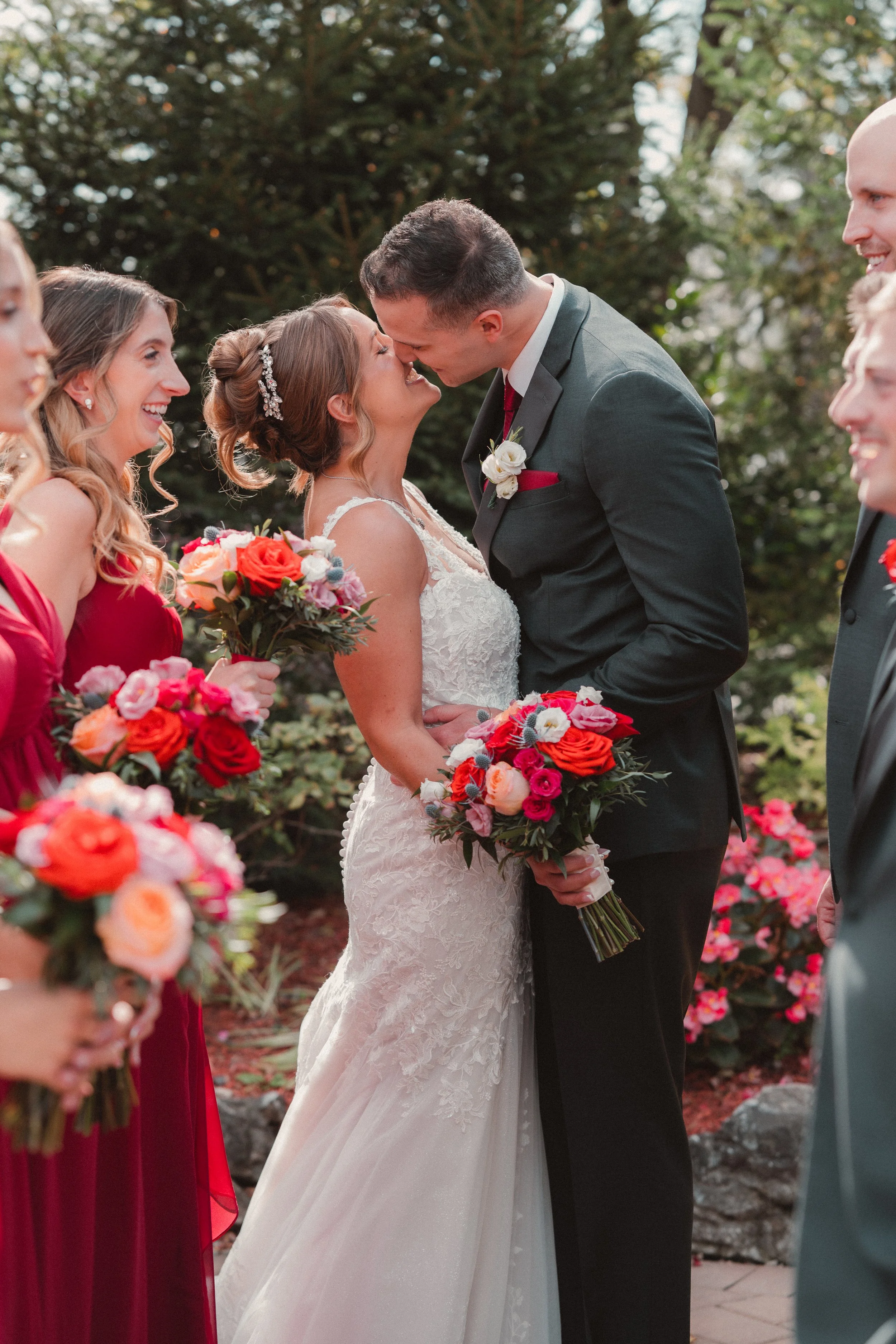 A wedding ceremony taking place outdoors in front of trees. The bride and groom are about to kiss, standing close to each other. The bride is in a white wedding dress holding a bouquet of flowers, and the groom is in a dark suit with a red tie and bo
