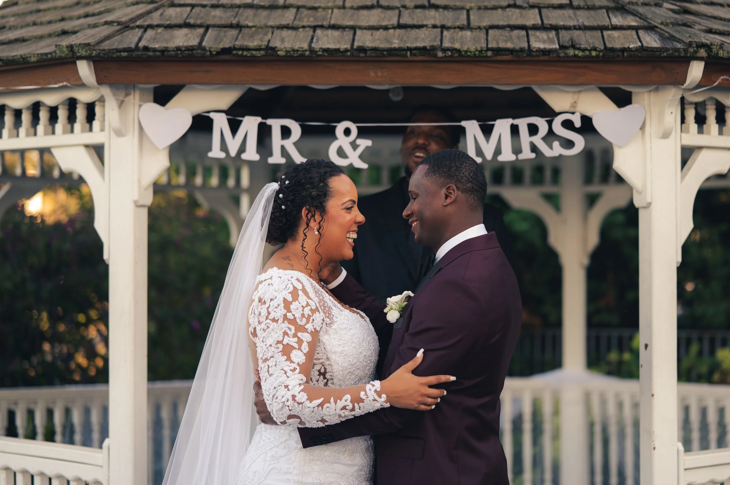 A bride and groom smiling and embracing during their wedding ceremony under a white gazebo decorated with a "MR & MRS" banner and heart-shaped ornaments.
