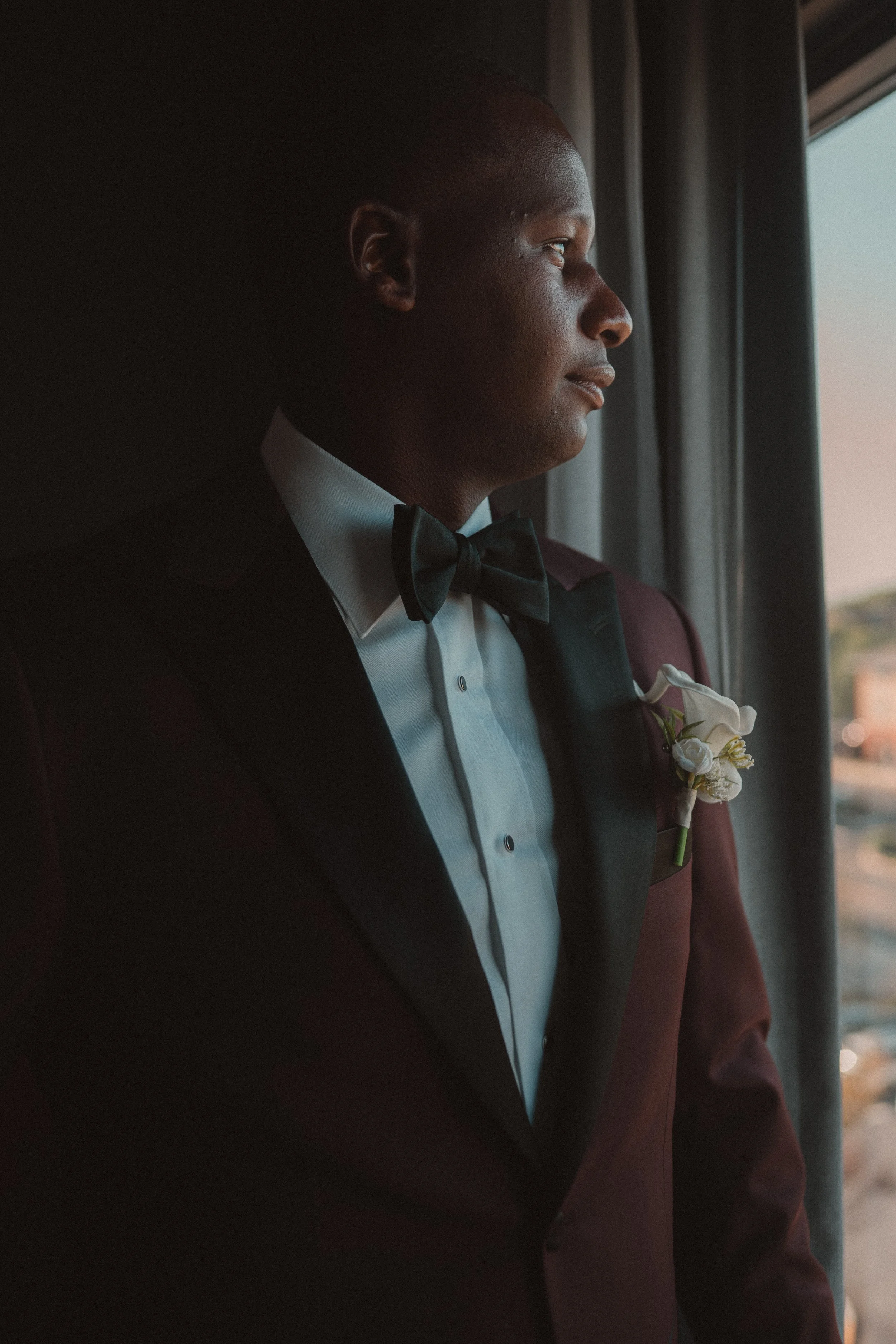 Portrait of a groom in a tuxedo with a boutonniere, looking out the window.