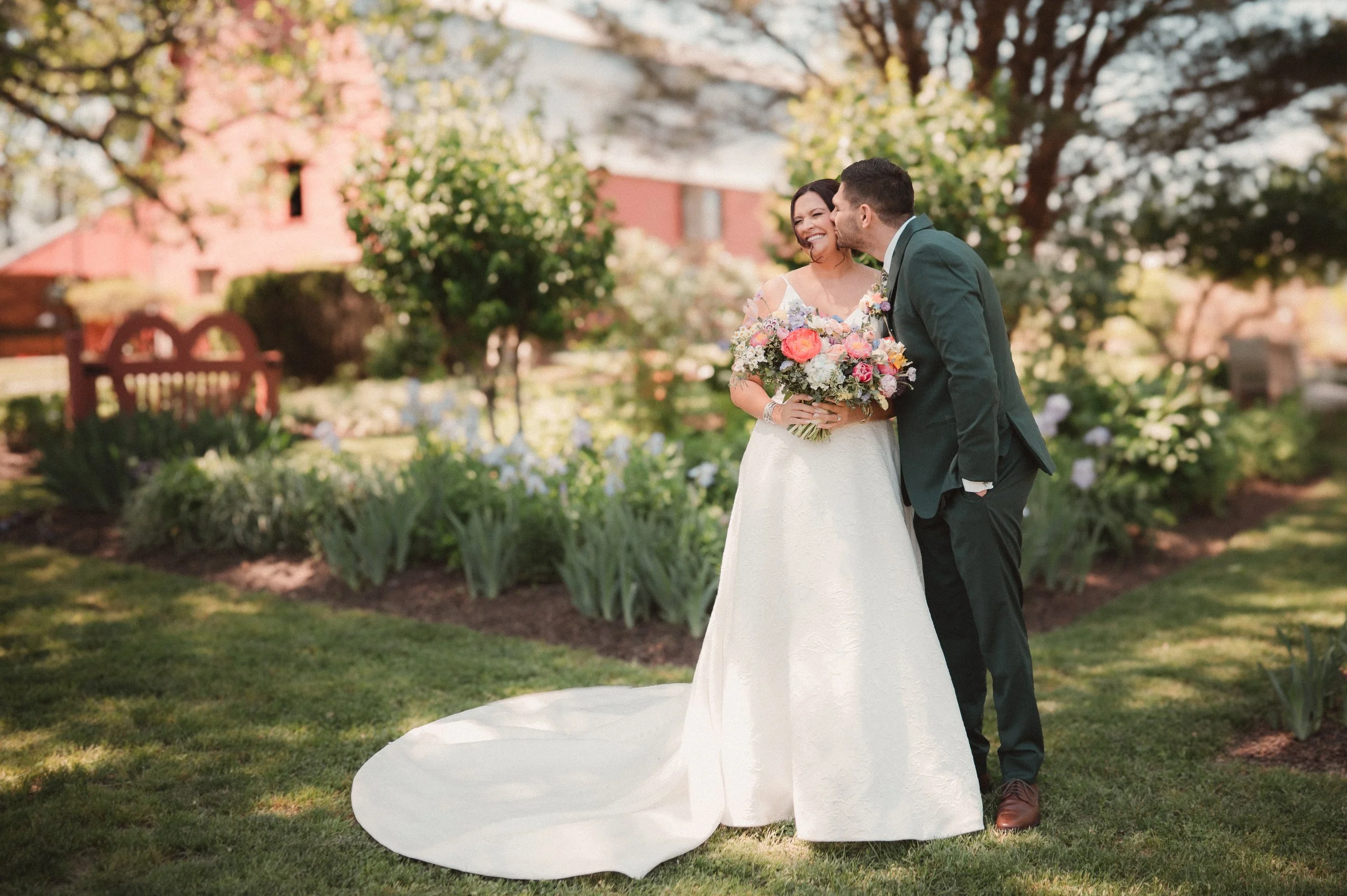 A bride in a white wedding dress holding a colorful bouquet, and a groom in a dark green suit sharing a kiss on the cheek in a garden with trees, flowers, and a red house in the background.