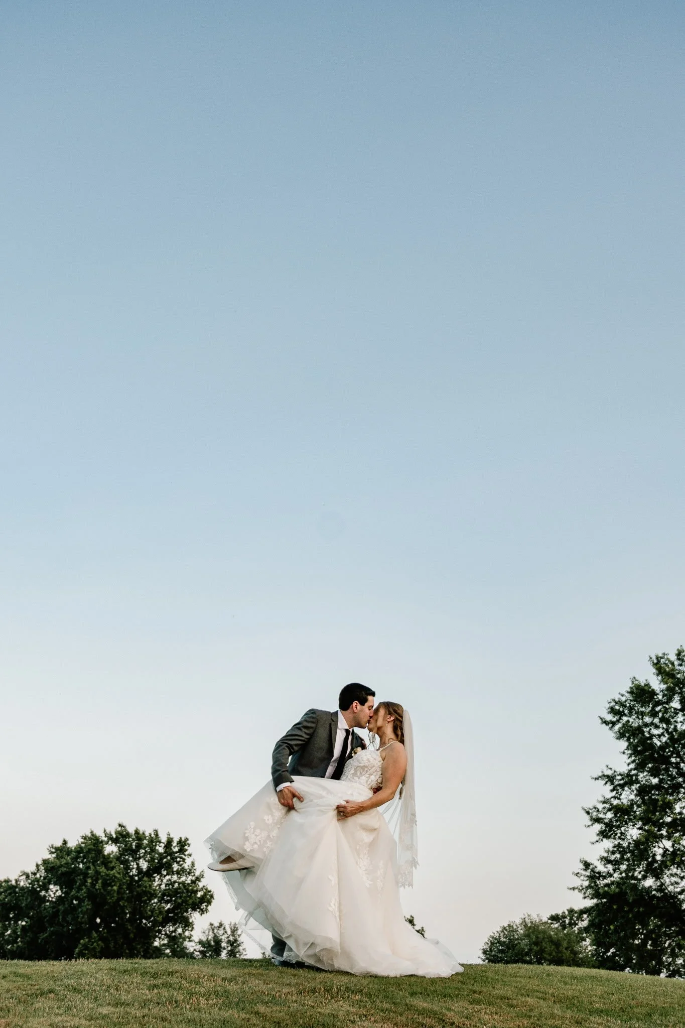 A newlywed couple sharing a kiss outdoors during sunset, with trees in the background.