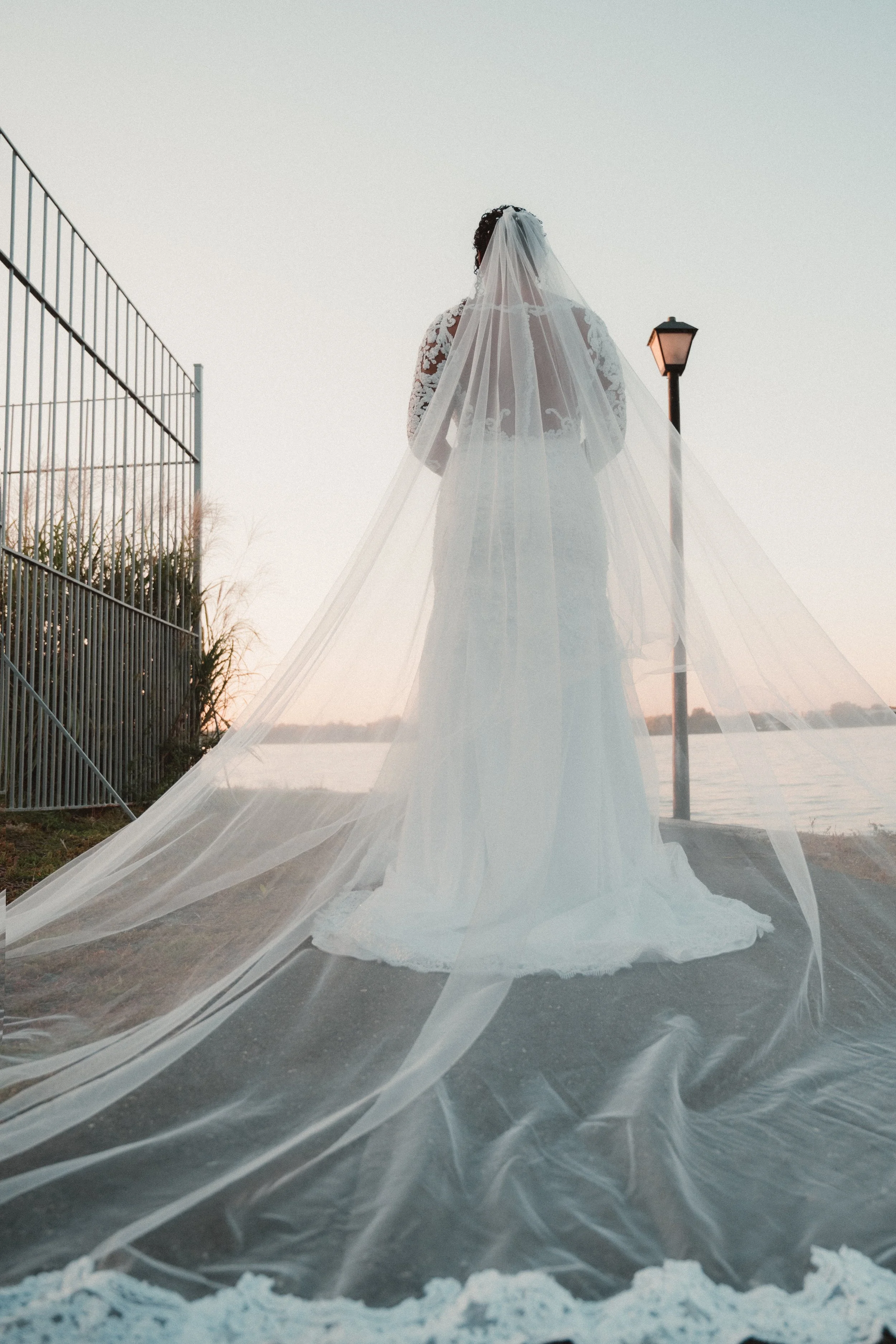 A bride in a wedding dress with lace details, standing outdoors near a body of water at sunset, wearing a long, flowing veil.