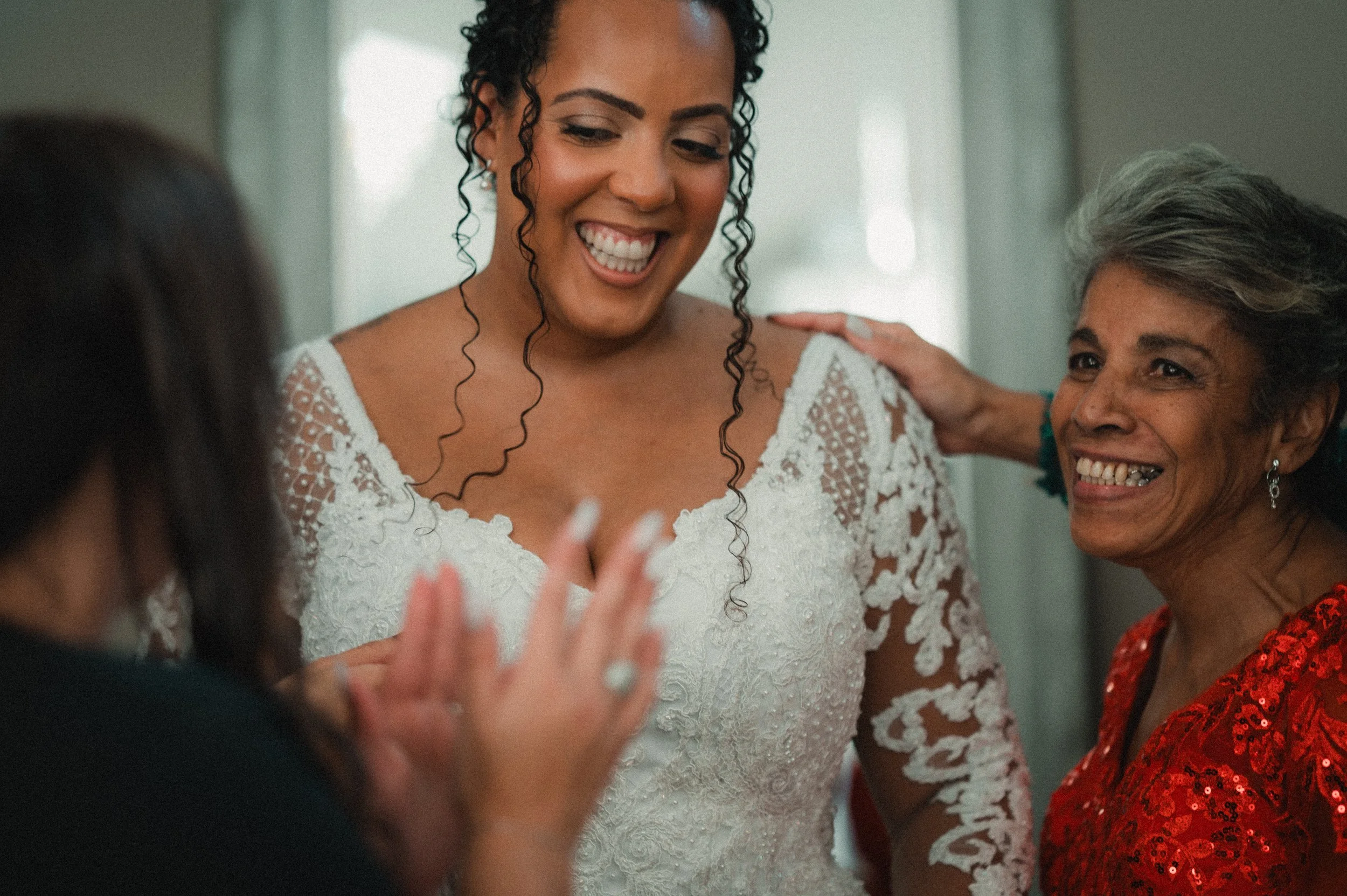 A bride in a white laced wedding dress smiling and talking with two women, one with gray hair in a red sequined dress and another woman with dark hair, in a bright indoor setting.
