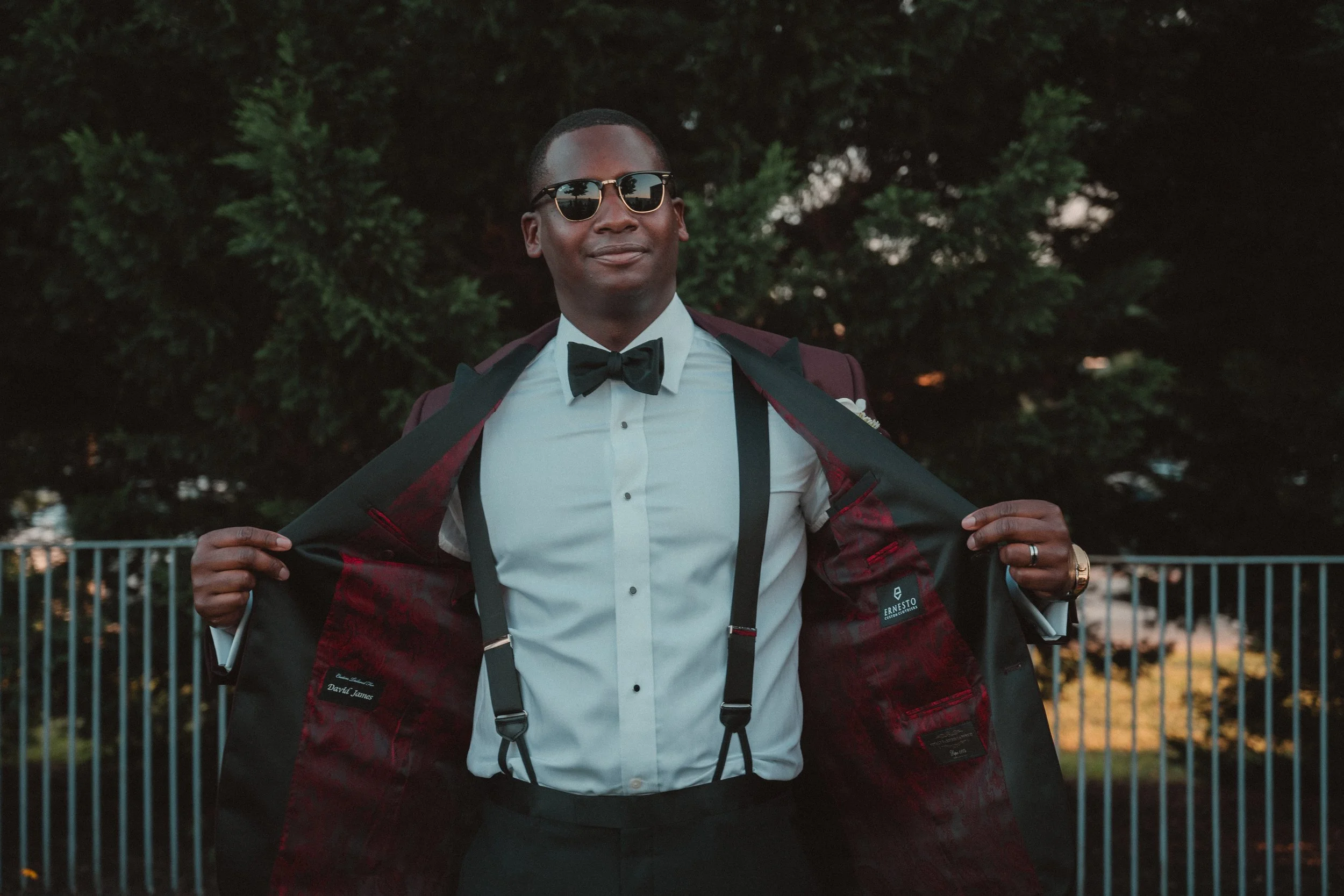 A man dressed in formal attire, wearing sunglasses, a bow tie, suspenders, and a tuxedo jacket, opening jacket to show its lining outdoors with trees in background.