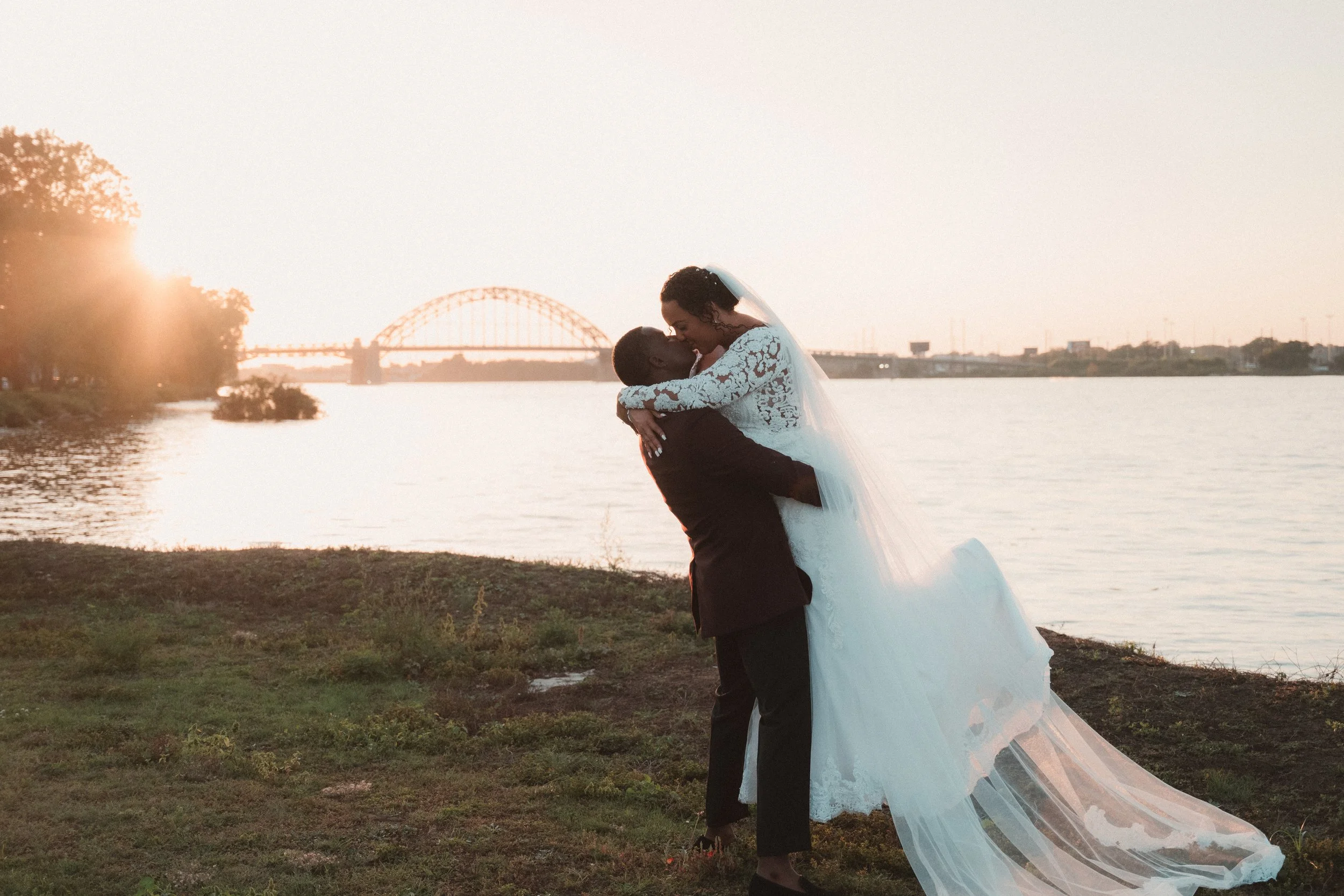 A bride and groom sharing a kiss by a river at sunset with a bridge in the background.