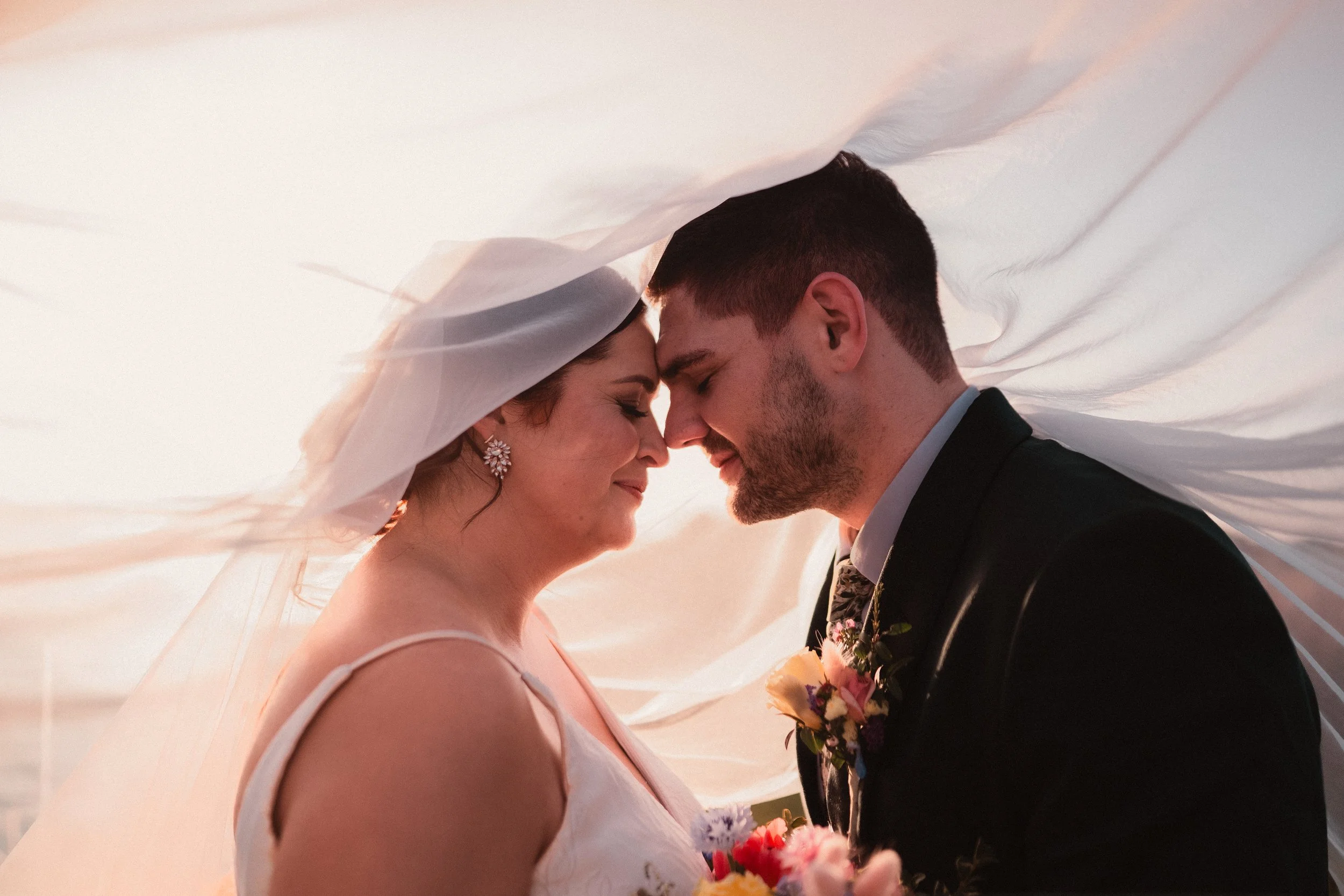 A bride and groom face each other with their foreheads touching, smiling softly in a wedding portrait, both under a flowing veil.