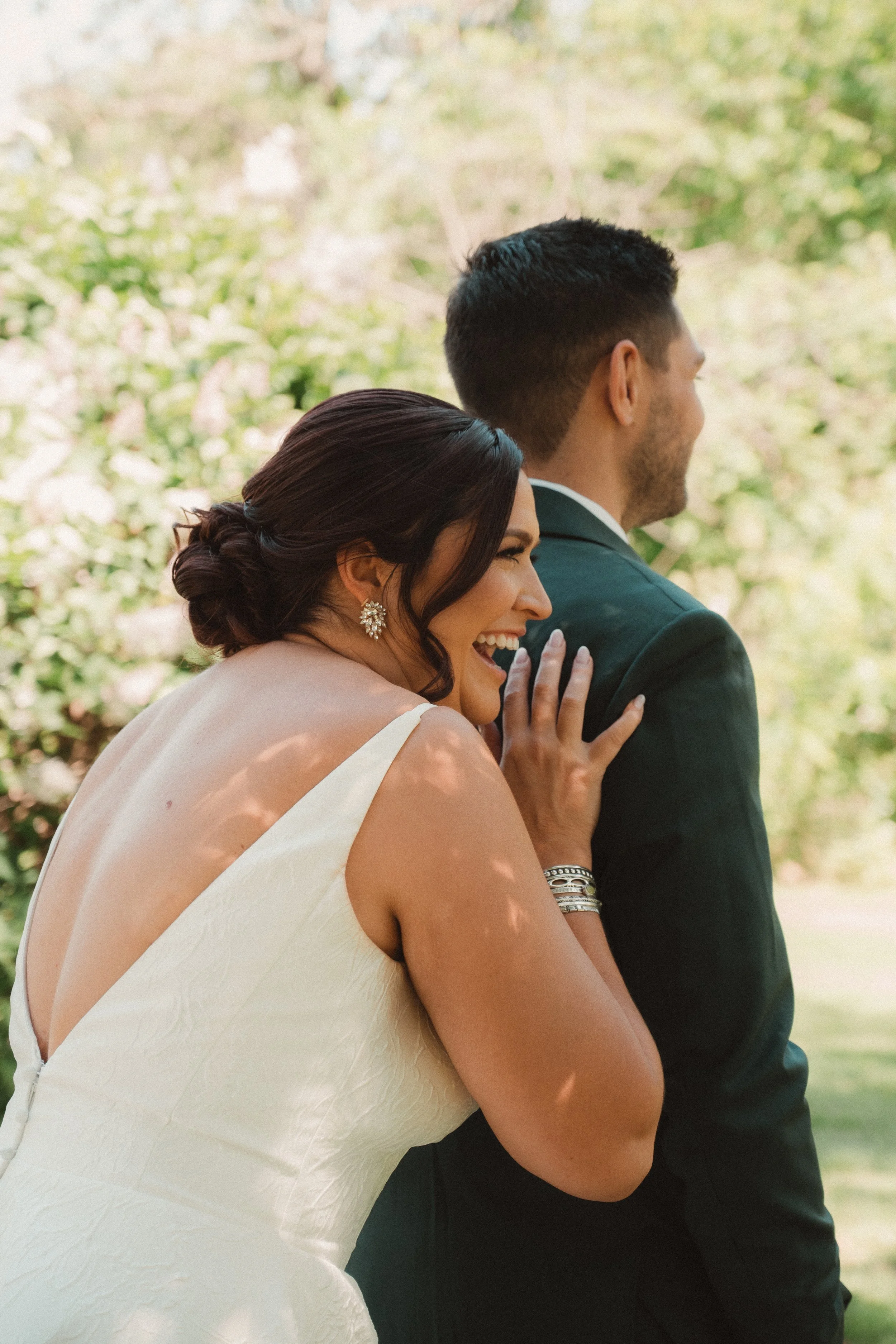 A bride and groom outdoors, the bride laughing and touching the groom's shoulder, surrounded by greenery.