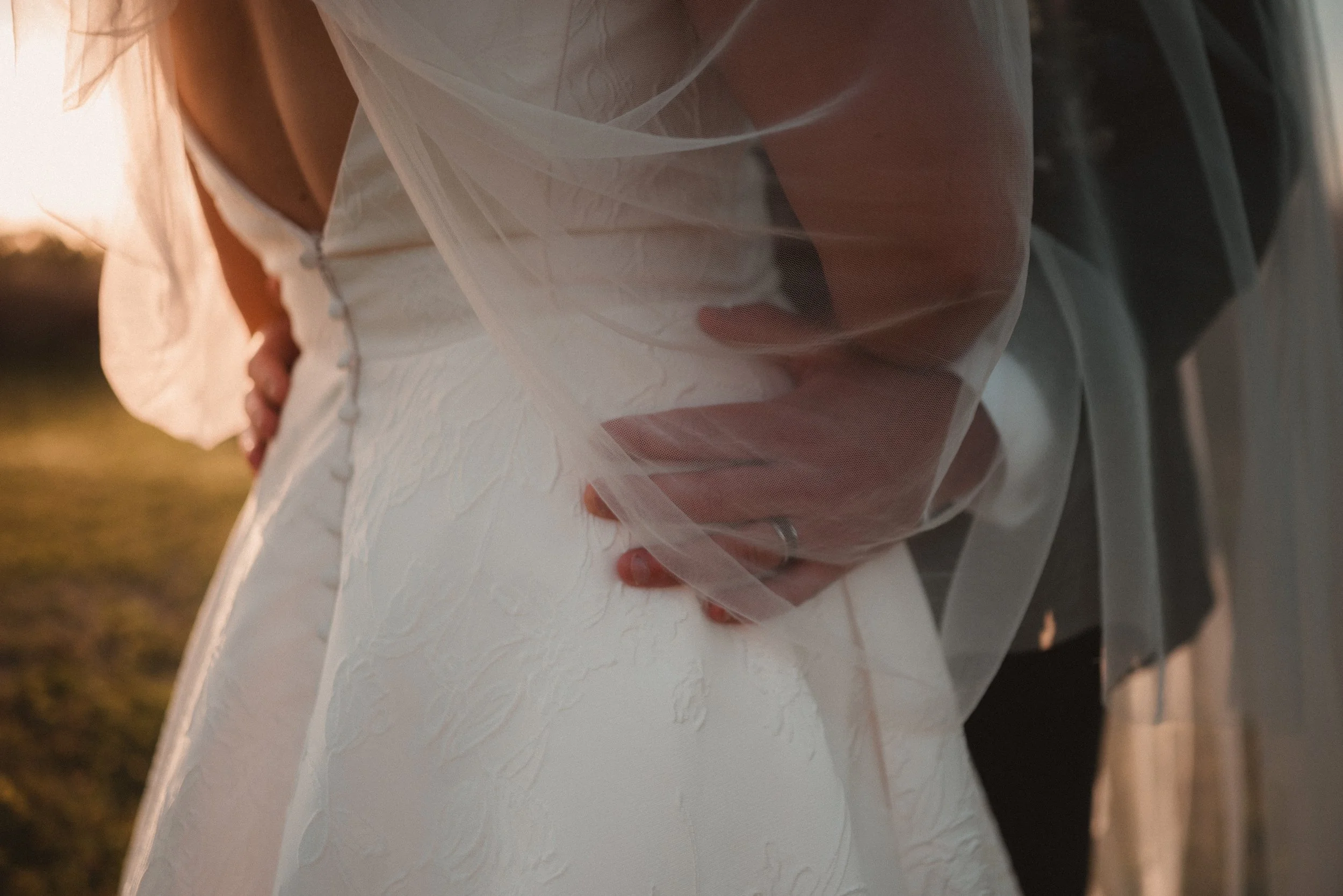 Close-up of a bride in a white wedding dress with lace details, lightly holding her hip, during sunset outdoors.
