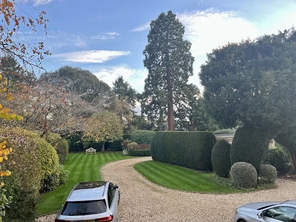 A gravel driveway curves through a lush garden with trimmed bushes and trees, a silver car parked in the foreground, and a cloudy sky overhead.