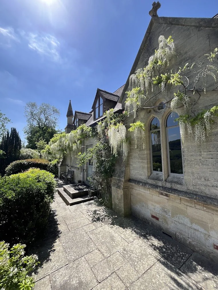 Stone building with arched windows and climbing white flowering vines, set against a bright blue sky with some clouds and lush greenery.