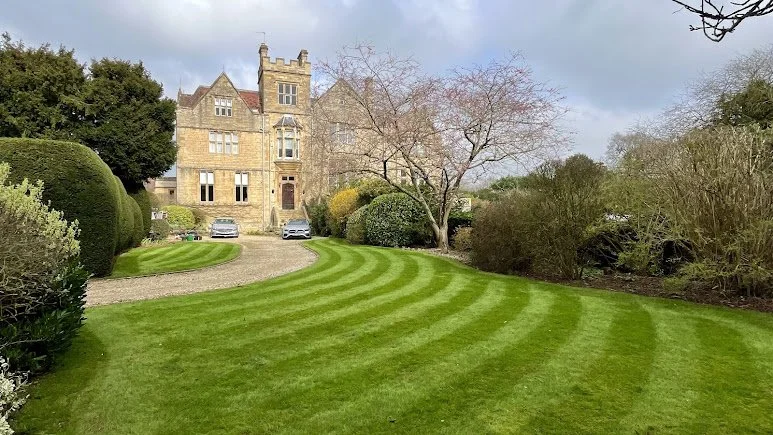 A large historic stone mansion with a well-maintained green lawn in front, featuring curved mowing lines, surrounded by trees and shrubs.