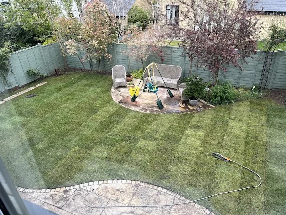 Backyard with a green lawn, a small patio with two chairs and a swing set, trees along the fence, and a garden hose on the grass.