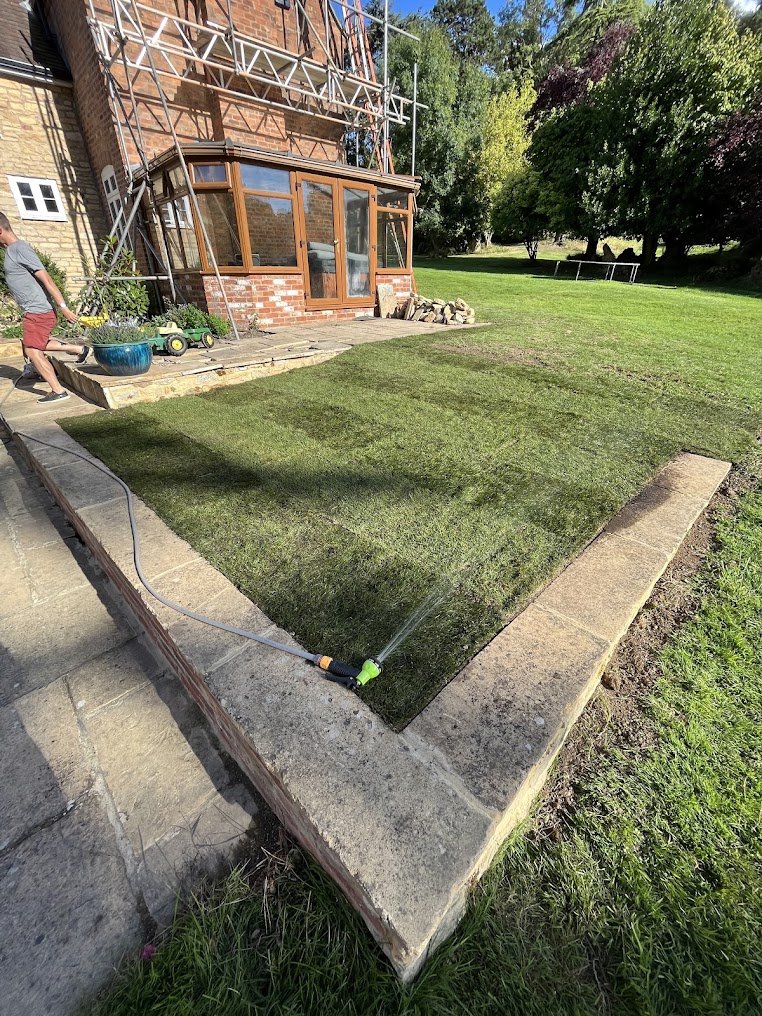 A person watering a freshly laid patch of grass in a backyard.